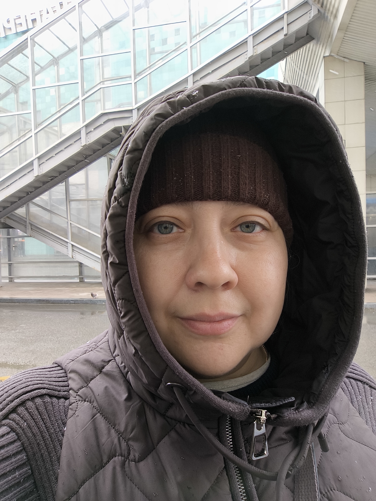 A medium, eye-level shot shows a woman wearing a brown knit hat and a dark-colored hooded jacket with water droplets. The jacket has a quilted pattern on the front and knit sleeves. Her hood is up, framing her face. She has fair skin and blue eyes.
Behind her, a metal staircase with glass panels leads to a building. The scene appears to be outside, and it is likely wet due to the droplets on the woman's jacket. Based on the architectural style and the presence of the Cyrillic script visible on the building in the upper left corner of the image, the location is Moscow, Russia. A small bird is present on the ground behind her.