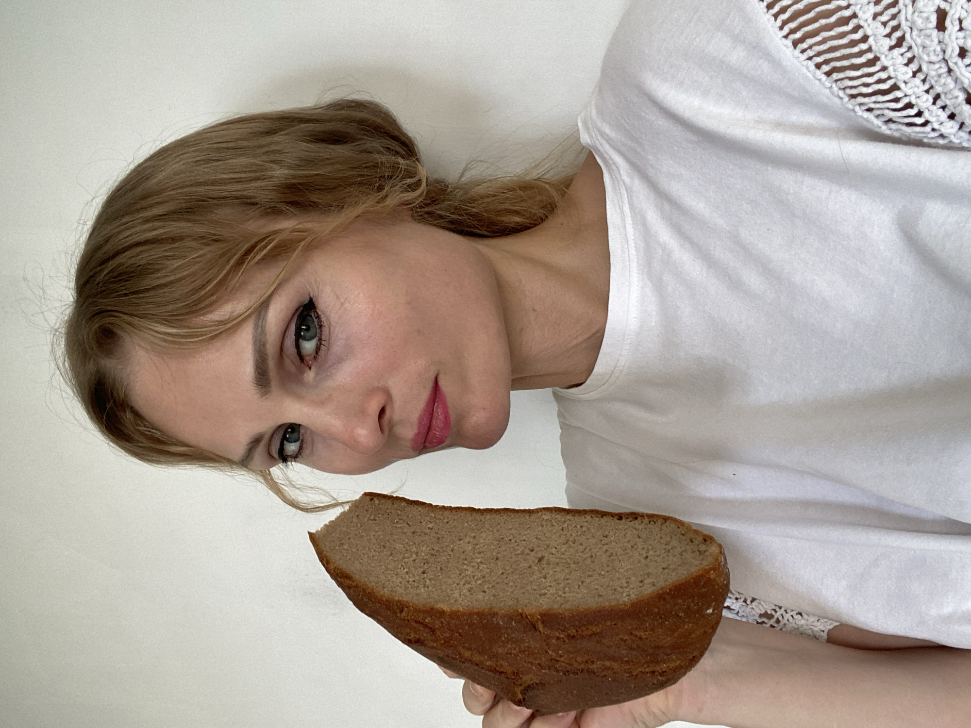 A fair-skinned woman with light brown hair pulled back holds a slice of dark bread to her face, her left eye in focus and her lips slightly parted. She is wearing a white t-shirt with crochet detailing on the sleeves and is positioned against a plain, white background. The lighting is even, creating no harsh shadows. The overall scene is a close-up portrait. The provided metadata indicates the image was taken in Міханавіцкі сельскі Савет, Belarus, however, the image itself offers no contextual clues to confirm or deny this location, as it is a simple indoor shot.