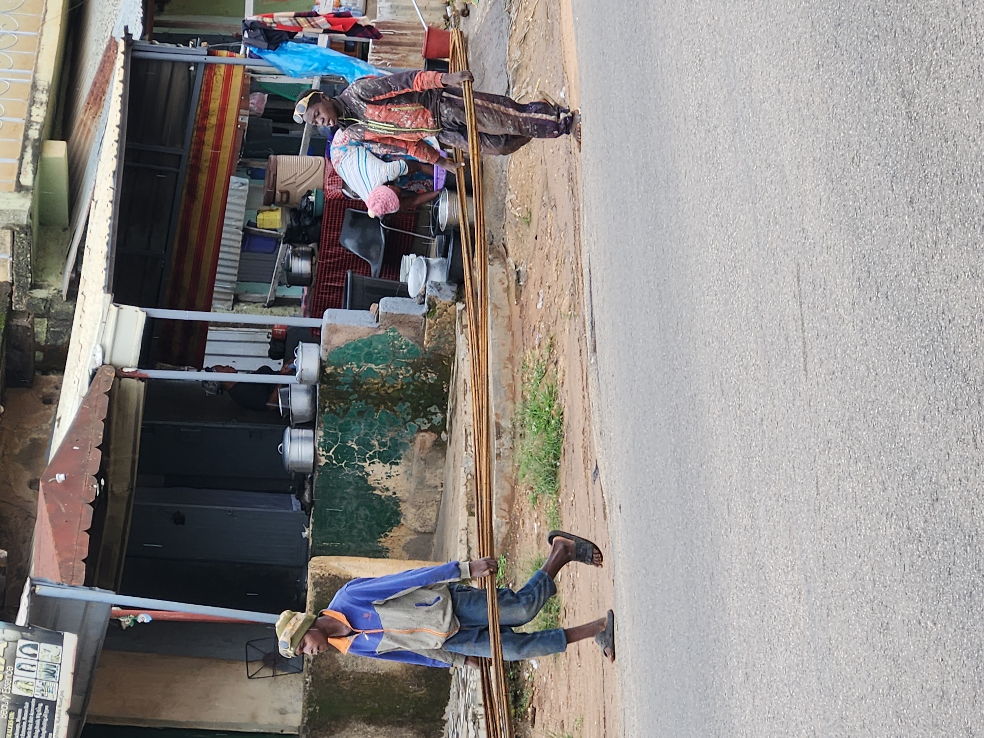 The image shows two people carrying metal rods along a street. The first person, on the left, is wearing a patterned jacket and pants and a cap. They are holding a bundle of metal rods horizontally across their body. Behind them, another person is partially visible, wearing a striped shirt and a pink hat. They appear to be interacting with items in a stall.
The second person, on the right, is walking along the side of the road, also carrying a bundle of metal rods. They are wearing a blue and grey jacket, blue jeans, a cap, and sandals. They are looking forward as they walk.
The scene appears to be an outdoor market or street in a developed area, with buildings and stalls lining the street. The ground is a mixture of dirt and pavement. There is some vegetation visible. The lighting suggests it is daytime, and the weather appears to be overcast or slightly hazy. There are no clear indicators of the specific time of day or weather conditions beyond this.
One building in the background has a sign that is partially visible. The text on the sign includes "Beauty Essence" and "Shoulders so". There are also images of hairstyles on the sign. The overall atmosphere seems to be one of daily life and commerce.
