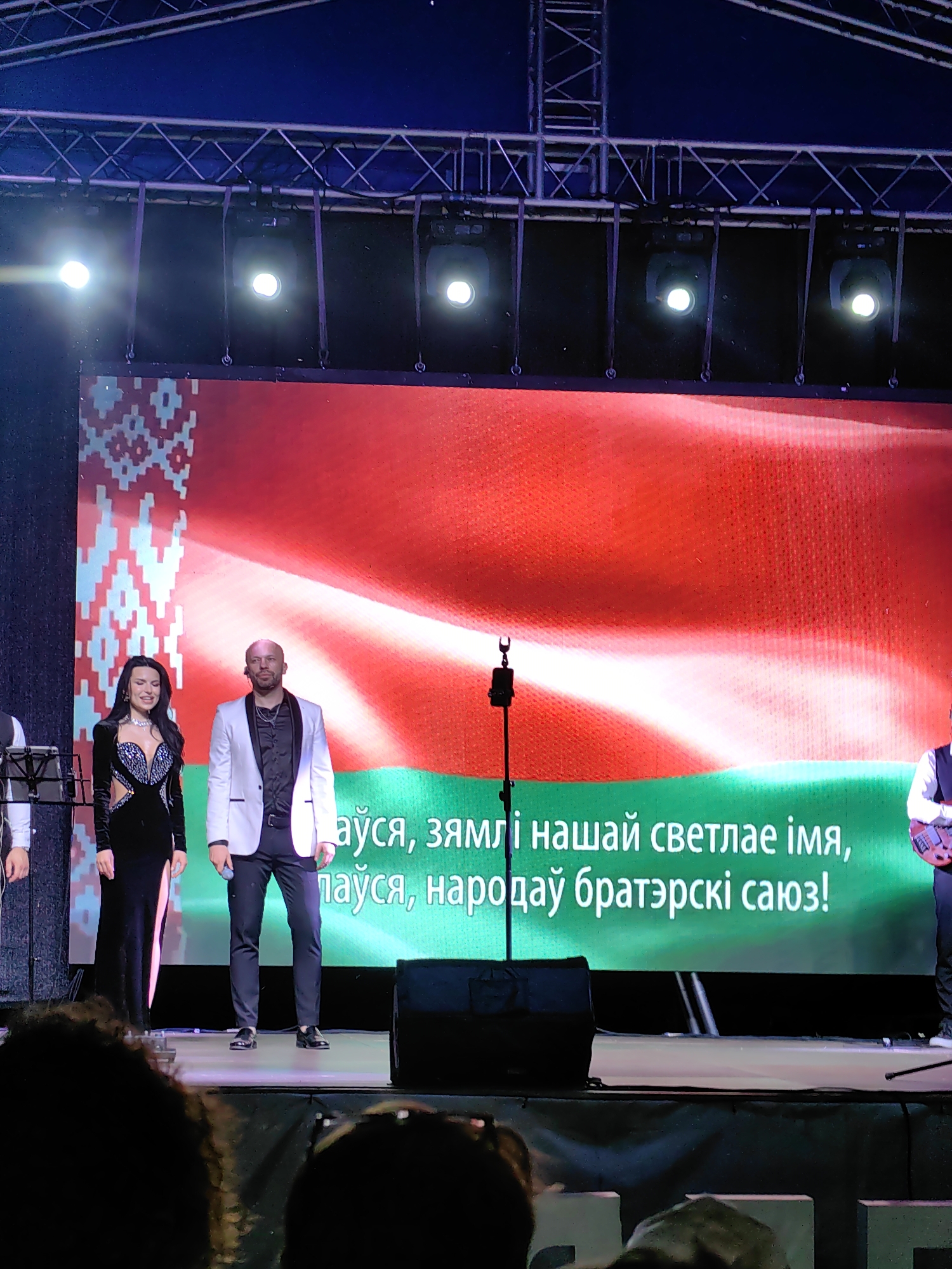 The image shows a stage performance in Myadzyel, Belarus. A woman in a black dress and a man in a white jacket and black shirt are standing on stage in front of a large LED screen displaying the Belarusian flag. The screen also shows text in Belarusian. There are stage lights above the performers, and speakers are visible on the stage. The audience is visible in the foreground. The overall scene suggests a concert or cultural event.