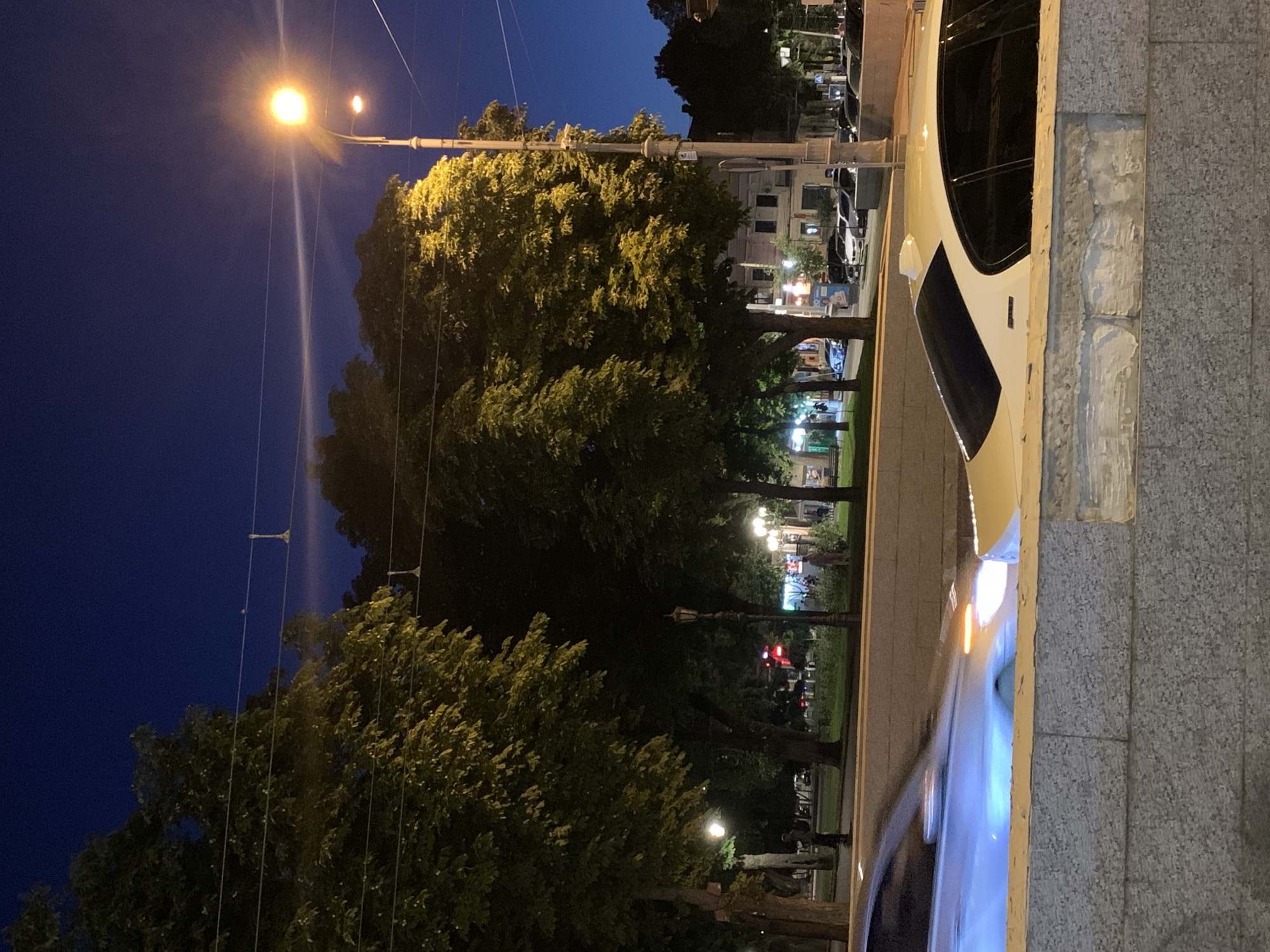 In the city of Odesa, Ukraine, at dusk, a street scene unfolds. The sky is a deep twilight blue, with a faint glow from the setting sun casting a light haze. Tall, leafy trees line a street, their foliage illuminated by a warm streetlamp positioned to the right of the frame. The streetlamp emits a bright yellow light, casting long shadows.
In the foreground, a white car with tinted windows is parked partially obscured by a grey stone wall. To the left, another car, possibly white or light blue, is captured in motion, its headlights blurred by the speed, creating streaks of light. The car's front lights are illuminated with orange and white light.
Behind the trees, buildings with visible windows and lights can be seen, suggesting a commercial or residential area. A vintage-style lamppost stands in the middle ground, adding to the urban ambiance. Several figures are visible in the background, walking or standing, their features indistinct in the low light. The overall impression is a tranquil evening scene in a European city.