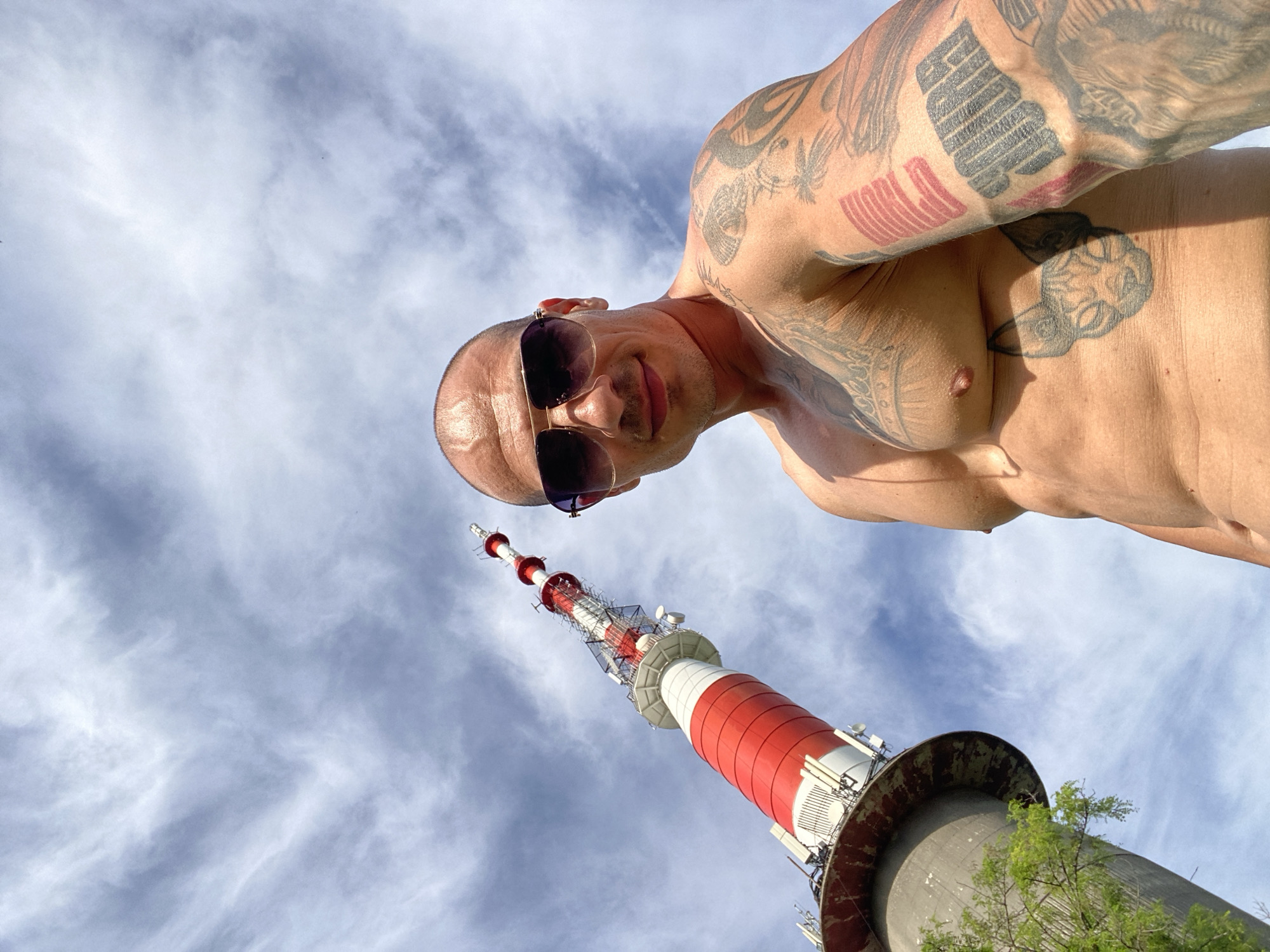 A low-angle, wide shot captures a man posing in front of the Pleven Television Tower. The man, who is shirtless and has extensive tattoos on his chest and arms, is wearing sunglasses. He is positioned to the right of the frame, looking towards the camera. To his left, the Pleven Television Tower rises dramatically against a sky filled with wispy clouds. The tower is characterized by its red and white striped pattern. The surrounding environment appears to be outdoors, with the base of the tower visible and some greenery on the left side of the frame.