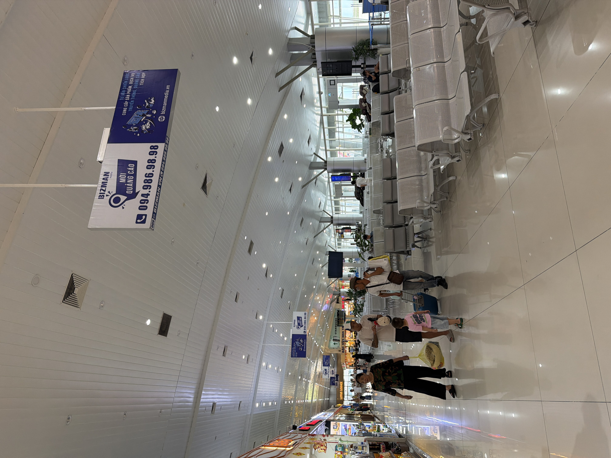 The image depicts the interior of a modern airport terminal in Cam Ranh Ward, Vietnam. The scene is brightly lit, with a high, white, slatted ceiling featuring recessed lighting and air vents. In the foreground, a polished, light-colored tiled floor reflects the overhead lights and the activity above.
Several people are visible walking through the terminal. On the left, a woman in dark clothing with a colorful patterned top carries a yellow bag. Beside her, a young boy in a pink t-shirt and shorts walks with two adults. A man in a light-colored polo shirt and a cap is in front of the boy, and another man in a white t-shirt and a cap, carrying a dark bag over his shoulder, walks slightly behind. They appear to be a family group. Further down the concourse, more people are seen walking and shopping in the brightly lit storefronts lining the left side of the terminal.
The right side of the terminal is characterized by rows of empty, light-colored metal seating arranged in parallel lines, suggesting a waiting area. Large windows on the far right offer a view of the outside, though the details are indistinct. A large, rectangular digital advertisement hangs from the ceiling in the middle of the concourse, displaying "BIZMAN" and contact information in Vietnamese. In the background, more seating areas and structural elements of the airport are visible. The overall impression is one of a busy, functional, and contemporary transportation hub.