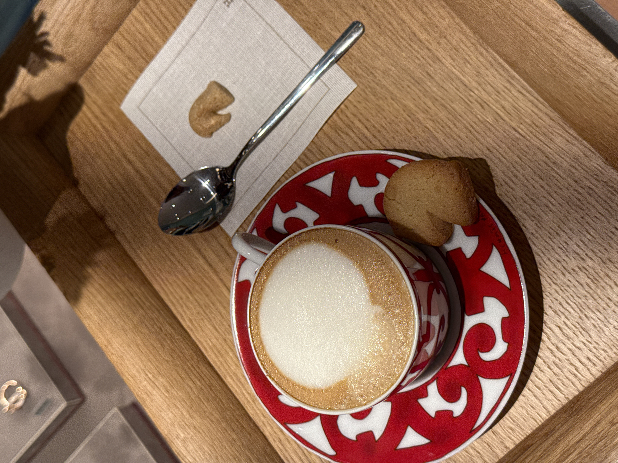 A close-up shot from a high angle depicts a cappuccino in a patterned red and white cup and saucer, alongside a napkin with a spoon and small cookie, all on a wooden tray. Another cookie is placed on the saucer, behind the mug. The scene appears to be indoors. The location in Zapopan, Mexico is not visually discernible from the image. No people are visible in the image.