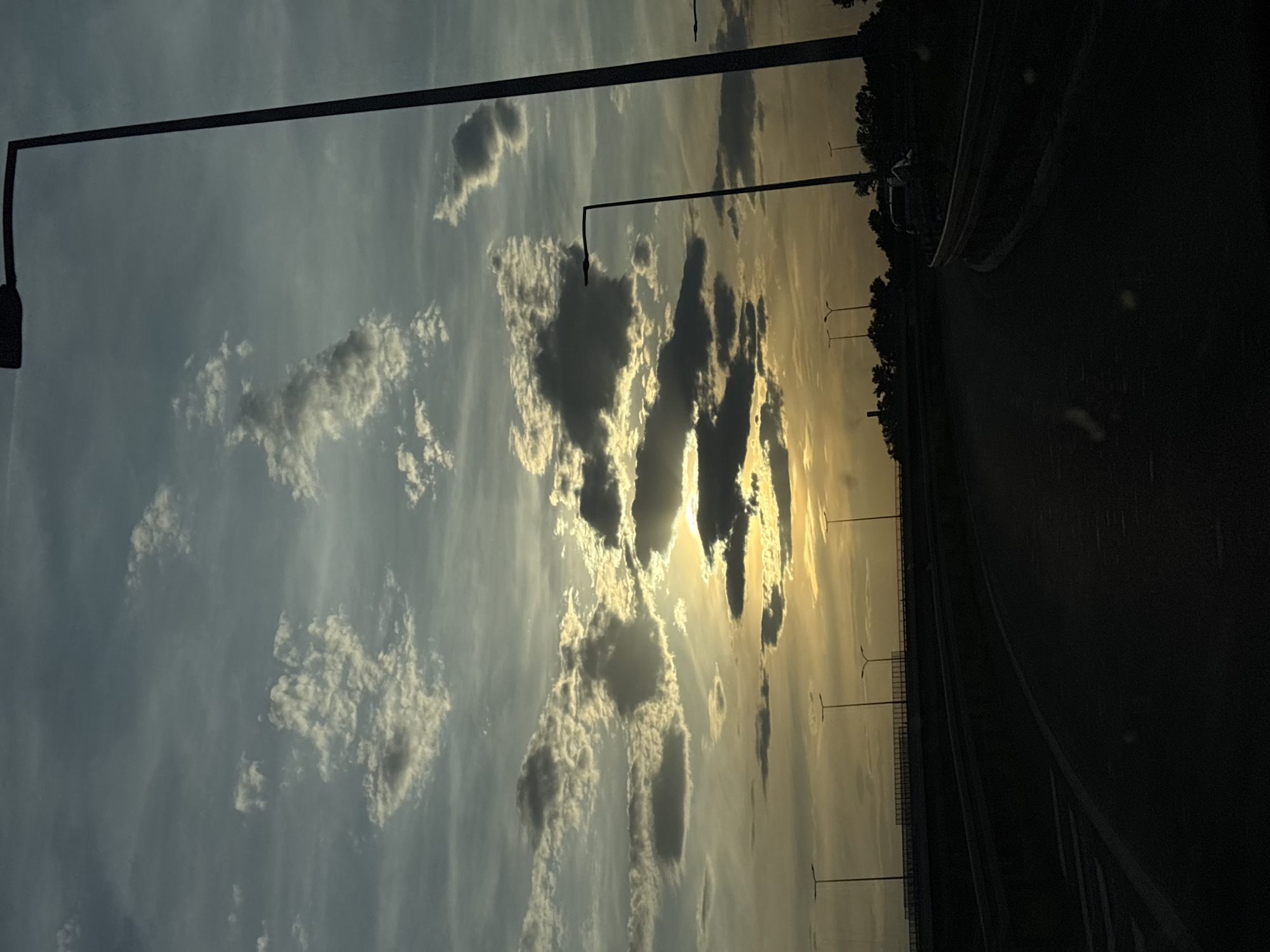 A landscape shot from a moving vehicle, likely a car, on a roadway in Obrenovac Urban Municipality, Serbia. The image is oriented sideways. The right side of the frame shows a dark asphalt road with a curved shoulder. The left side displays a sky filled with cumulus clouds, varying from white to dark gray, illuminated by the setting sun that casts golden rays. In the distance, evenly spaced streetlights stand along the road's edge. Overhead powerlines are visible against the sky. The shot appears to be taken during the sunset.