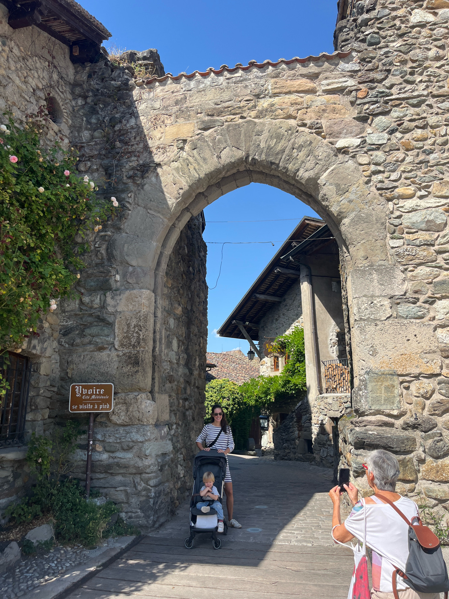 In the medieval town of Yvoire, France, a cobblestone path leads through an ancient stone archway. A woman with sunglasses and a striped dress pushes a stroller containing a young child. To the right of the arch, an older woman with gray hair takes a photo with her phone. A sign on the left wall reads "Yvoire Cité Médiévale se visite à pied" (Yvoire Medieval City to be visited on foot). Lush green foliage with pink roses climbs the stone walls, and the clear blue sky suggests a sunny day.
