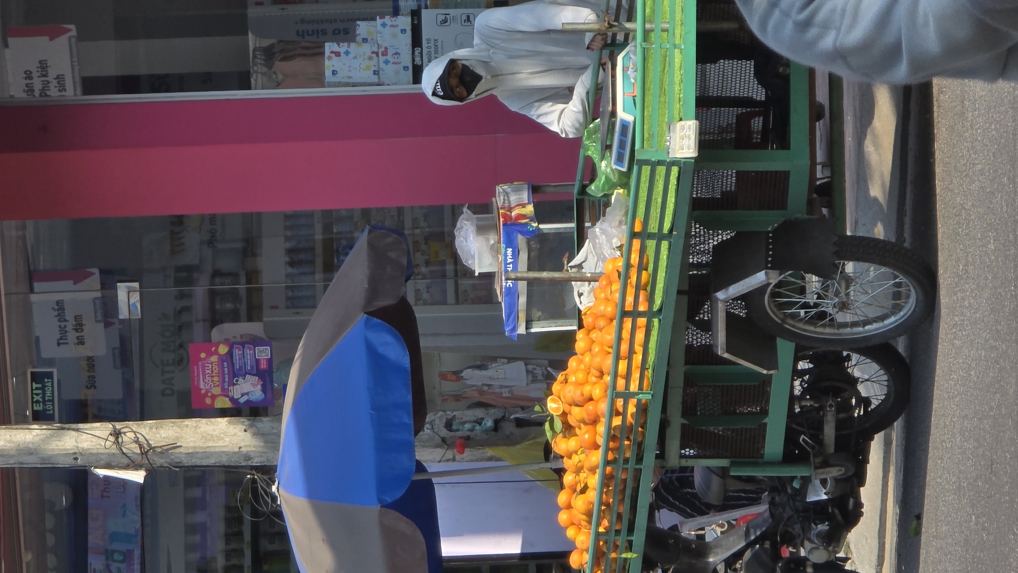The image shows a man in a white hooded jacket and black face mask operating a mobile orange vendor's cart. The cart is green and loaded with a large quantity of oranges. The cart is also equipped with a small weighing scale. Adjacent to the cart is a large umbrella. The background shows a storefront with signage written in Vietnamese. Signs on the storefront mention items like "Thực phẩm ăn dặm" (Infant Foods), "Sữa nước" (Milk), "DATE MOI," and "Jần áo Phụ kiện sinh" (Infant Clothes and Accessories). Based on the language and products displayed, the location is likely Bắc Nha Trang, Vietnam. A sidewalk and road are visible on the right side of the cart.
