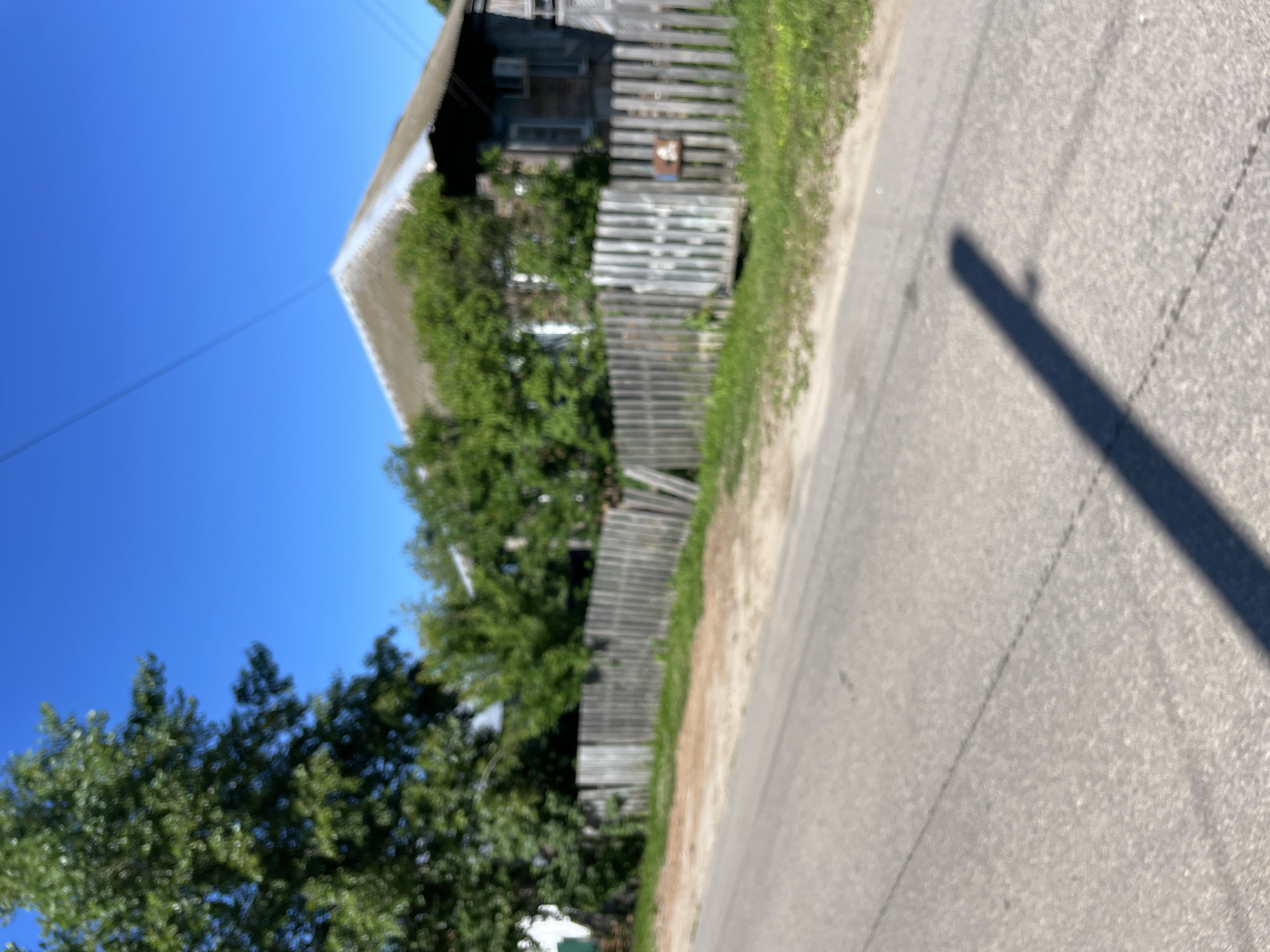 The image depicts a street scene in the village of Krasnye Baki, Russia. A weathered wooden fence runs along the side of a paved road, with a two-story wooden house visible behind it. The house appears to be older, with a metal roof and visible windows. Lush green trees and foliage surround the property. A shadow of what appears to be a utility pole is cast across the road. The sky is clear and blue, suggesting a sunny day. There are no people or animals visible in the frame.