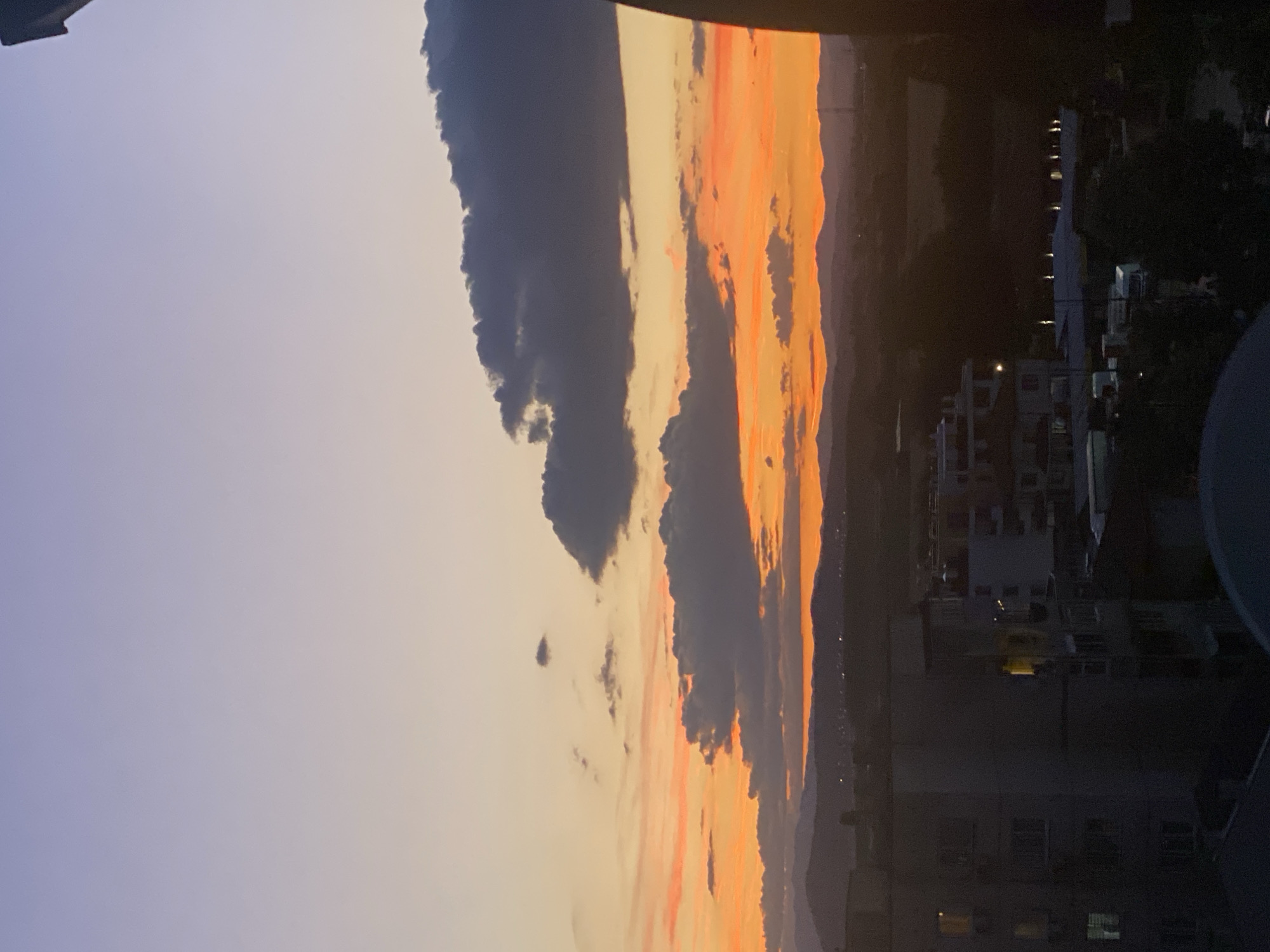 This image depicts a sunset over a city, likely Pazardzhik, Bulgaria, as indicated by the prompt. The sky transitions from a pale lavender at the top to vibrant oranges and yellows closer to the horizon, indicating the sun has recently set or is setting. Several dark, heavy clouds are scattered across the sky, partially obscuring the colorful hues.
In the foreground, the silhouette of buildings is visible against the twilight sky. The buildings appear to be multi-story residential structures, with some windows illuminated, suggesting indoor activity. The lower portion of the image shows a darker, more detailed view of these buildings, with trees and other urban elements discernible in the shadows. In the distance, a range of mountains forms a dark silhouette against the colorful sky, with faint lights of a settlement visible along their base. The overall atmosphere is tranquil, capturing a moment of urban transition from day to night.