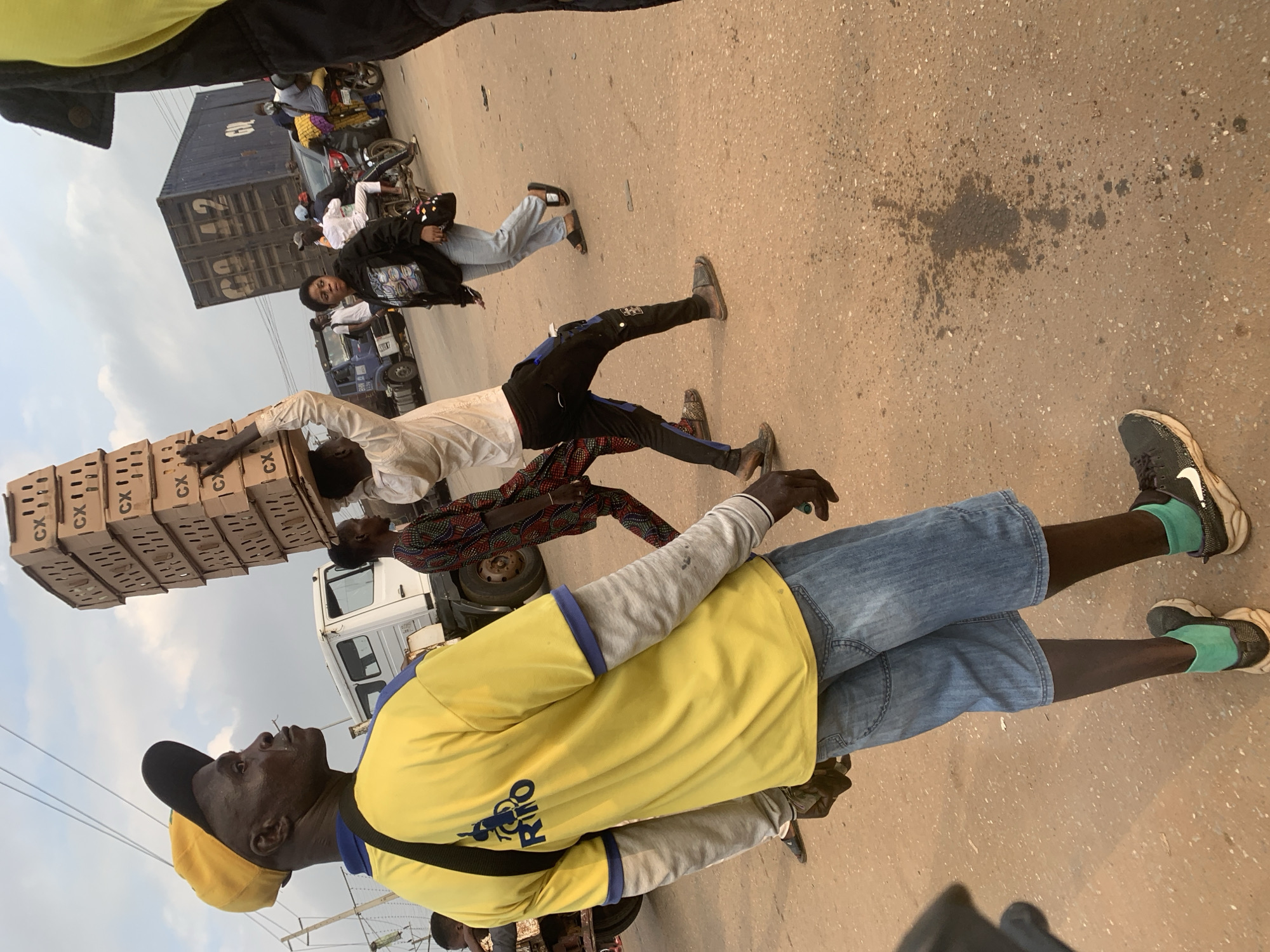 The image depicts a busy street scene in Shagamu, Nigeria, under an overcast sky. In the foreground, a man wearing a yellow t-shirt, blue denim shorts, and a yellow cap is observed from behind, facing to the left. He has a black strap across his chest. To his right, another man carries a tall stack of cardboard boxes on his head. Behind them, a white truck is parked. Further in the background, a large blue shipping container is visible, along with several motorcycles, cars, and pedestrians. The ground is paved with a reddish-brown surface, showing some scattered debris and a dark liquid stain. The overall atmosphere suggests a commercial or market area with ongoing activity.