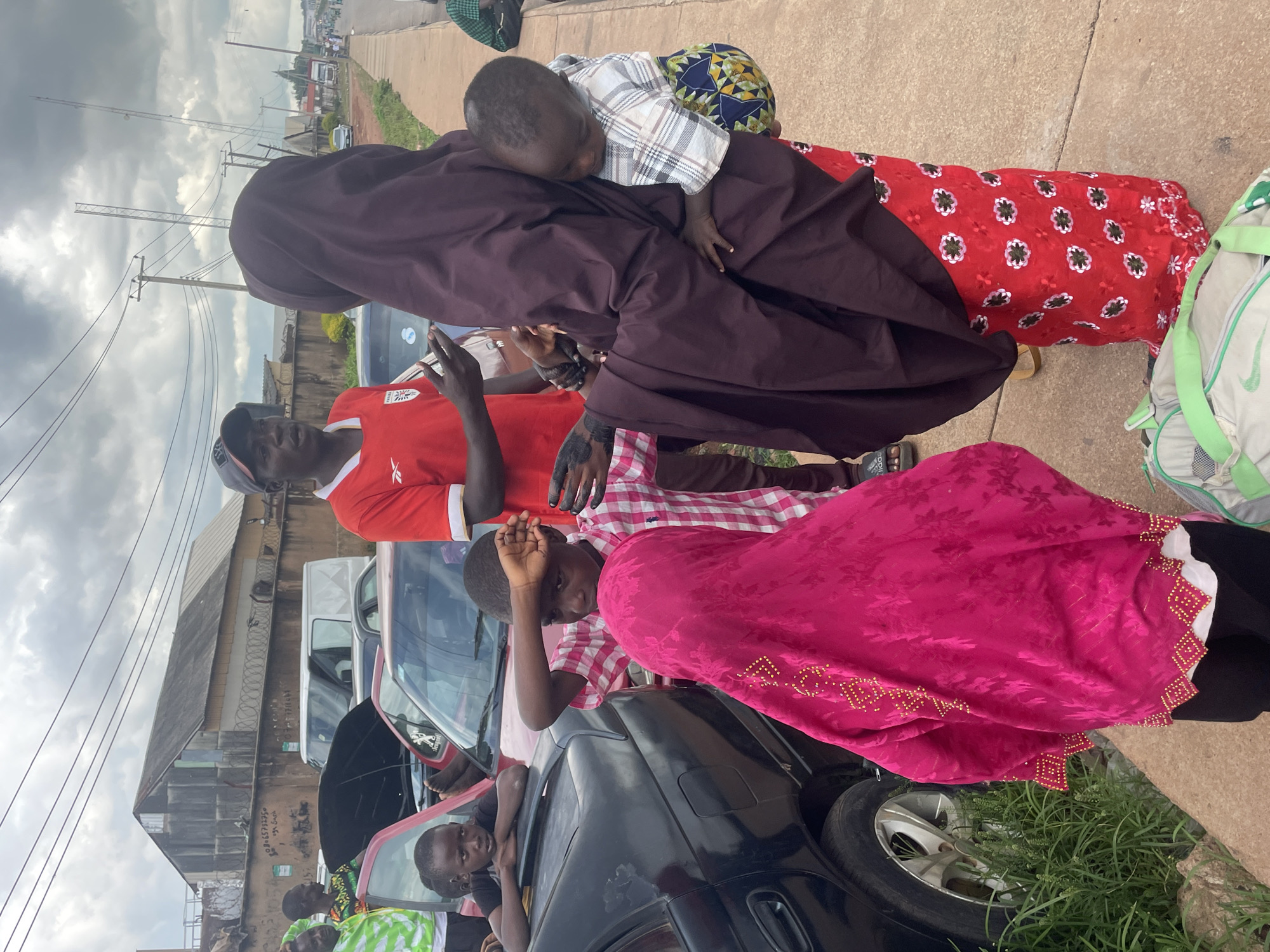 In Jos, Nigeria, on a cloudy day, a group of people stand on a sidewalk next to parked cars. In the foreground, a woman in a dark hijab and a vibrant red floral dress is holding a young child. To her left, a child in a bright pink hijab looks down. To the right of the woman, a man in a red shirt and a baseball cap is talking and gesturing with his hands. Behind them, another child in a checkered shirt is leaning against a black car. Further back, several cars are parked, some with hoods open, and a white van is visible. In the background, there are buildings, power lines, and traffic on a road. A green backpack lies on the sidewalk in the foreground.