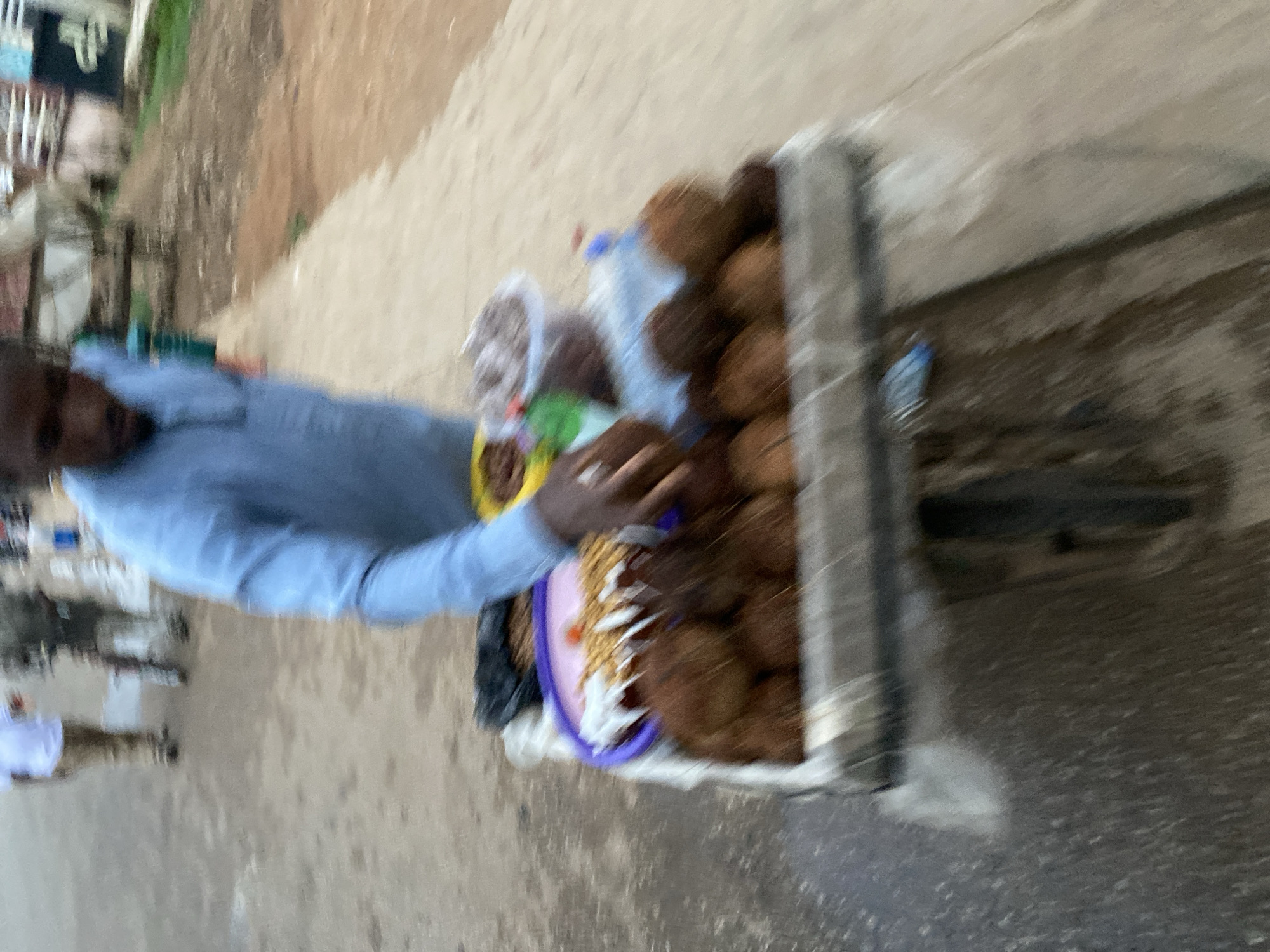 A vendor stands next to a cart filled with coconuts on a street in Kuru Dungus, Nigeria. He wears a blue shirt and appears to be handling other goods on the cart. Other people are visible in the background. The scene is an urban environment.