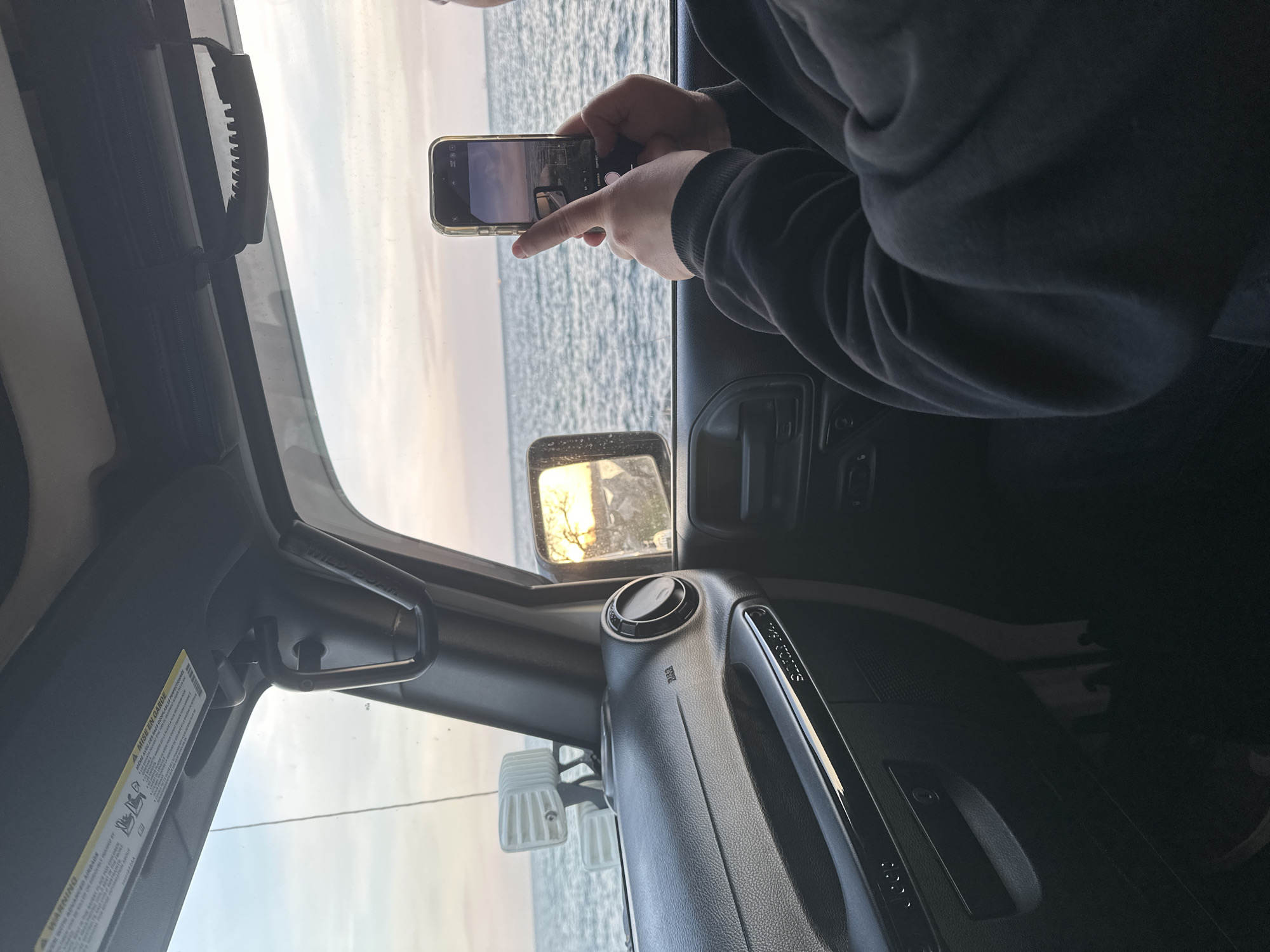 A person in a black sweatshirt takes a picture with their smartphone from inside a car, possibly a Jeep Wrangler, towards what appears to be the Black Sea near Varna, Bulgaria. The sea and horizon are visible in the smartphone's display and through the car windows. The car interior includes a dashboard, grab handle, side mirror reflecting sunlight on trees, and a sun visor with a warning label written in English and French. The focus is on the person using their phone to capture the seascape.