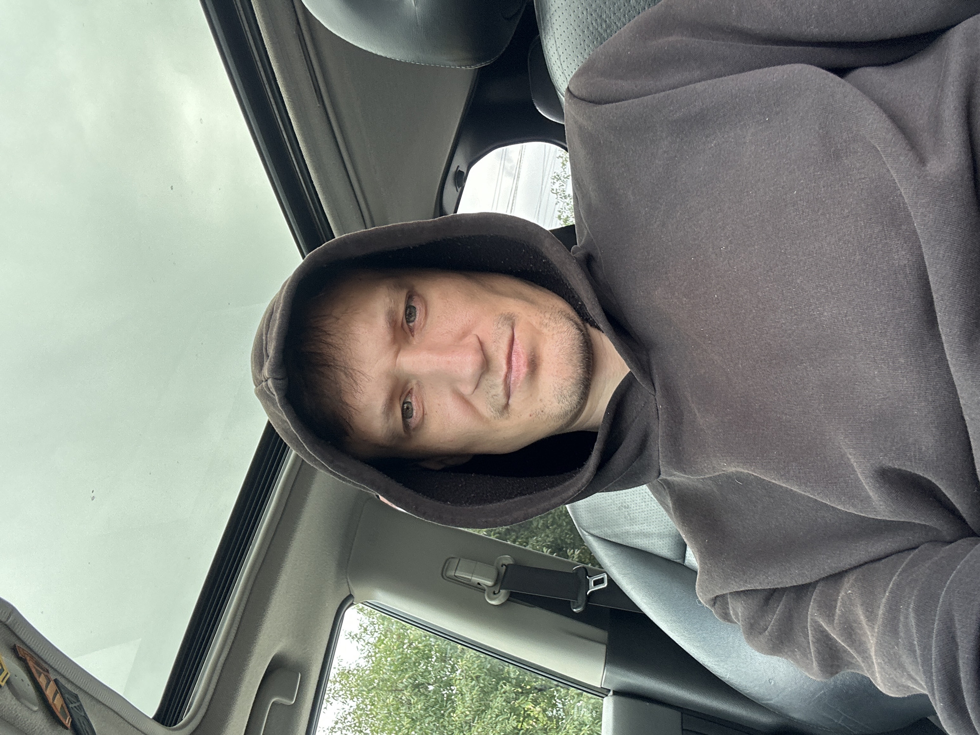 A man is sitting in the driver's seat of a car, wearing a dark hoodie. He is looking directly at the camera. The car has a sunroof that is open. The interior of the car is visible, including a black leather seat and a seatbelt. Outside the car, trees and greenery can be seen through the open sunroof and the car window. The sky appears overcast. The location is identified as Makiv, Ukraine.