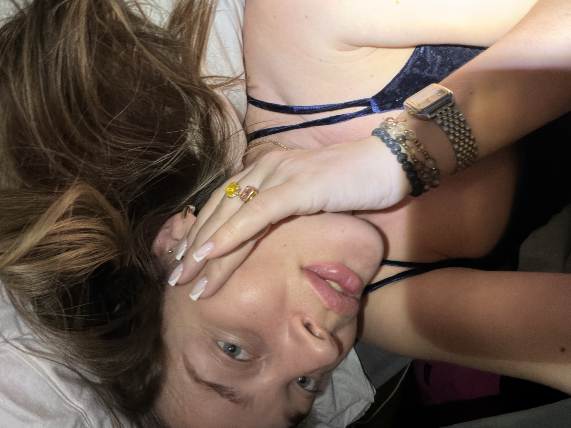 A woman lies on a white pillow, partially covering her face with her right hand. She has light skin, blue eyes, and brown hair. Her nails are manicured with a French tip. On her right hand, she wears several rings with colored stones. Her left arm is bent, and on her wrist, she wears a beaded bracelet and a watch with a metal band. She is wearing a dark-colored garment with thin straps. The environment appears to be indoors. There is no indication of the location being Kazan, Russia.