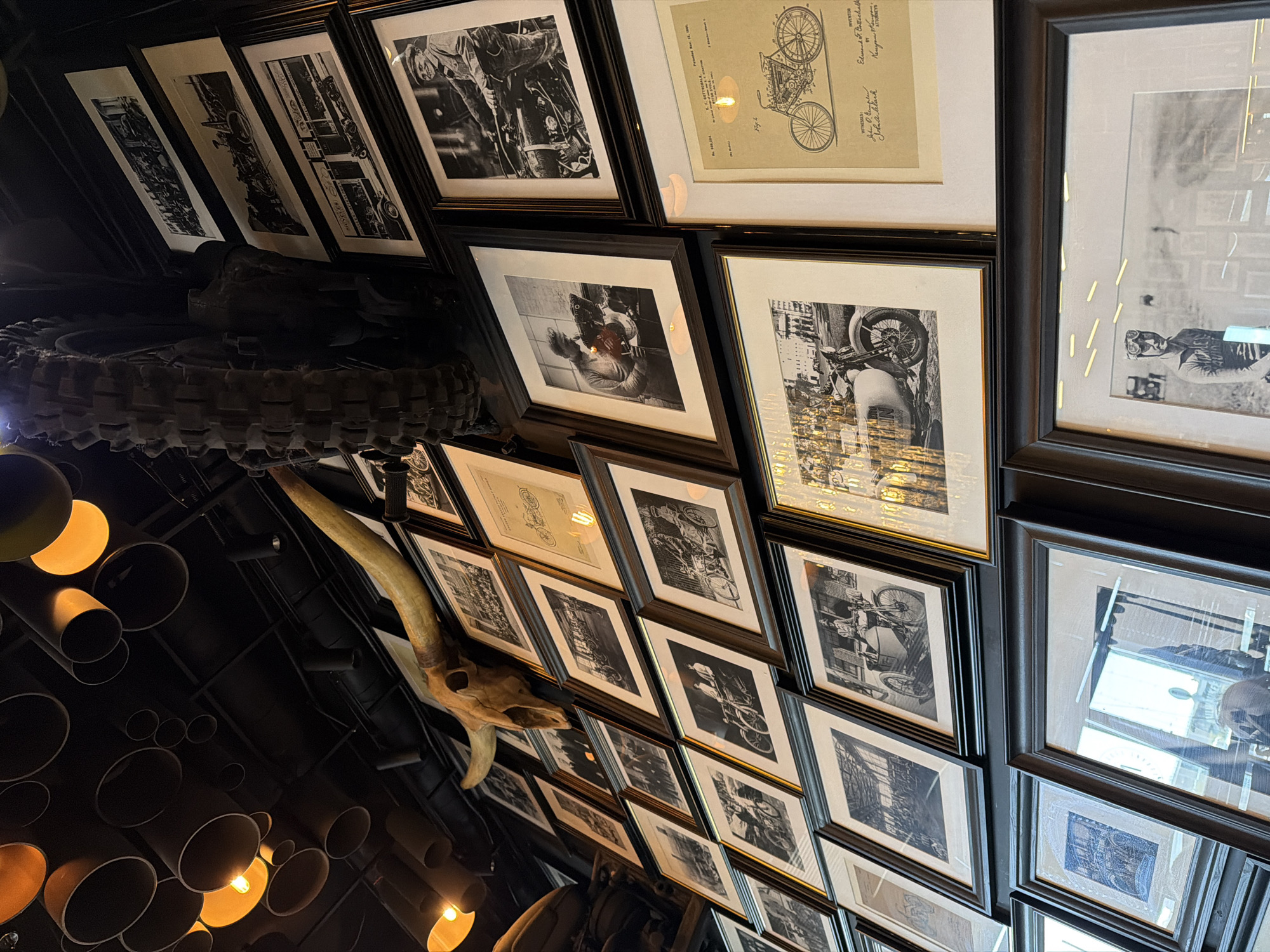 The image depicts the interior of a business establishment. The ceiling is decorated with numerous black-framed black and white pictures arranged in a grid. A large tire and part of a motorcycle are suspended from the ceiling, along with a skull with horns. The interior is dimly lit with yellow light fixtures hanging from the ceiling. The location could potentially be Saint Petersburg, Russia.
