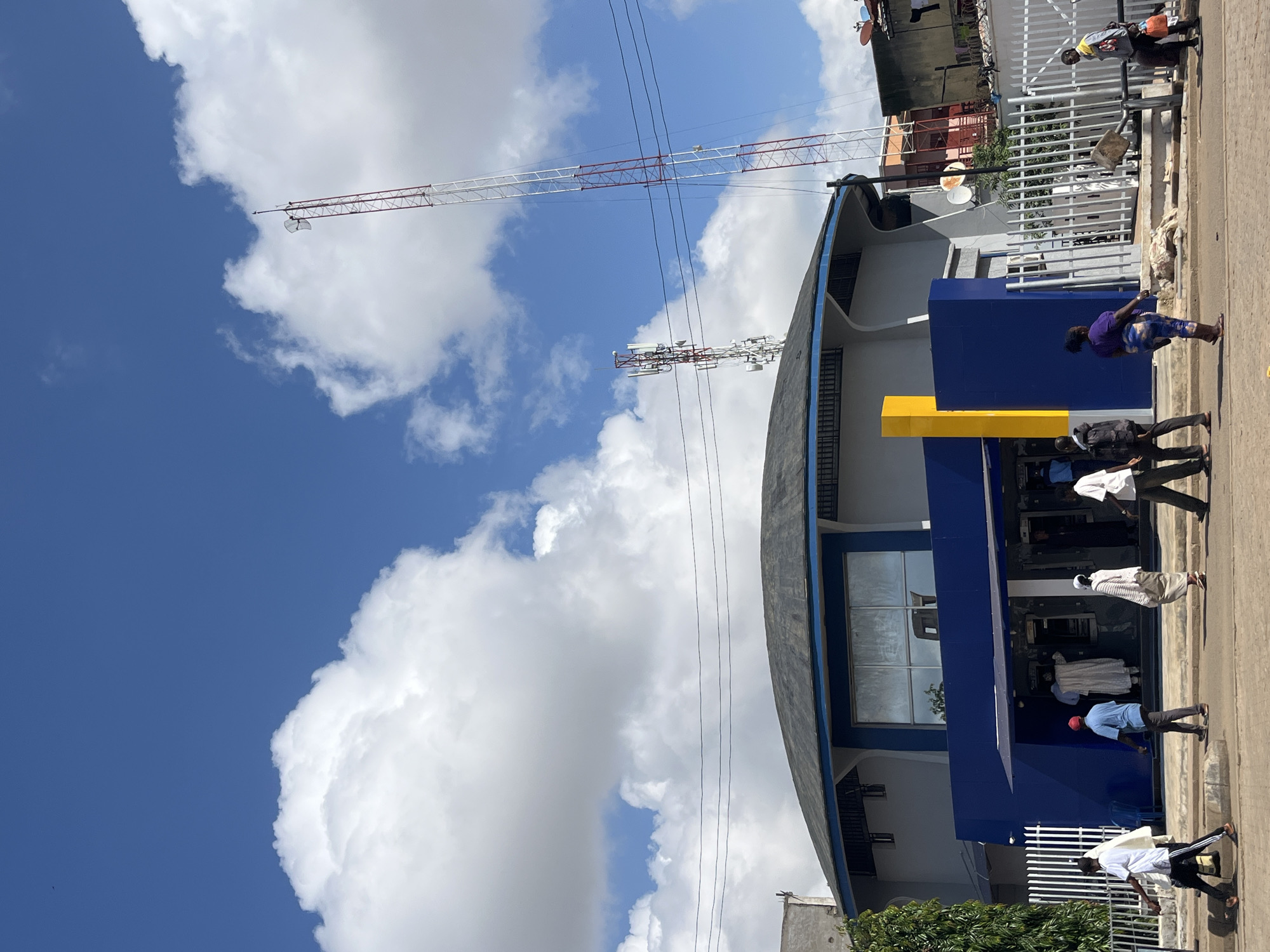This image depicts a street scene in Jos, Nigeria, on a sunny day with scattered clouds. The foreground features a curved blue building with large windows, a bright yellow architectural accent, and a black awning. Several people are visible on the sidewalk in front of the building, some appearing to be entering or exiting. One woman in a purple top and patterned leggings stands near the yellow accent, while others in white shirts and dark pants are further down the sidewalk.
In the background, a large cell tower or radio mast with red and white sections dominates the sky. Another, smaller tower with antennae is also visible. The buildings behind the blue structure appear to be multi-story and urban in character, with some showing signs of wear. The overall impression is of an active commercial or public area. The time of day appears to be midday, given the bright sunlight. There is no visible text in the image.