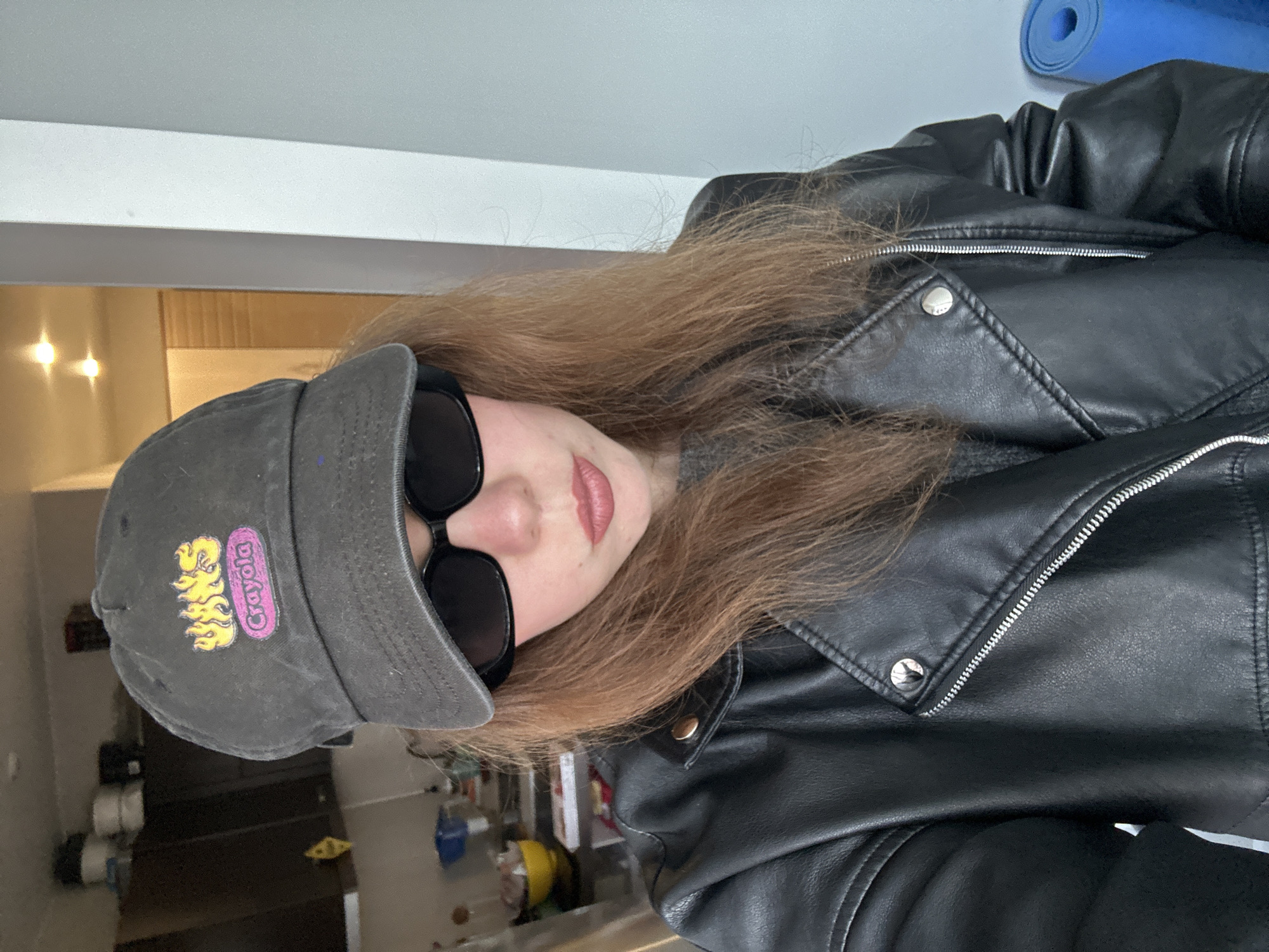 A person is wearing a black leather jacket, dark sunglasses, and a gray cap with a Crayola logo. The person is indoors. In the background are a blue yoga mat, a white wall, and a glimpse into another room with shelves or storage units. There is no indication of this picture being taken in the city of Krasnoyarsk, Russia.