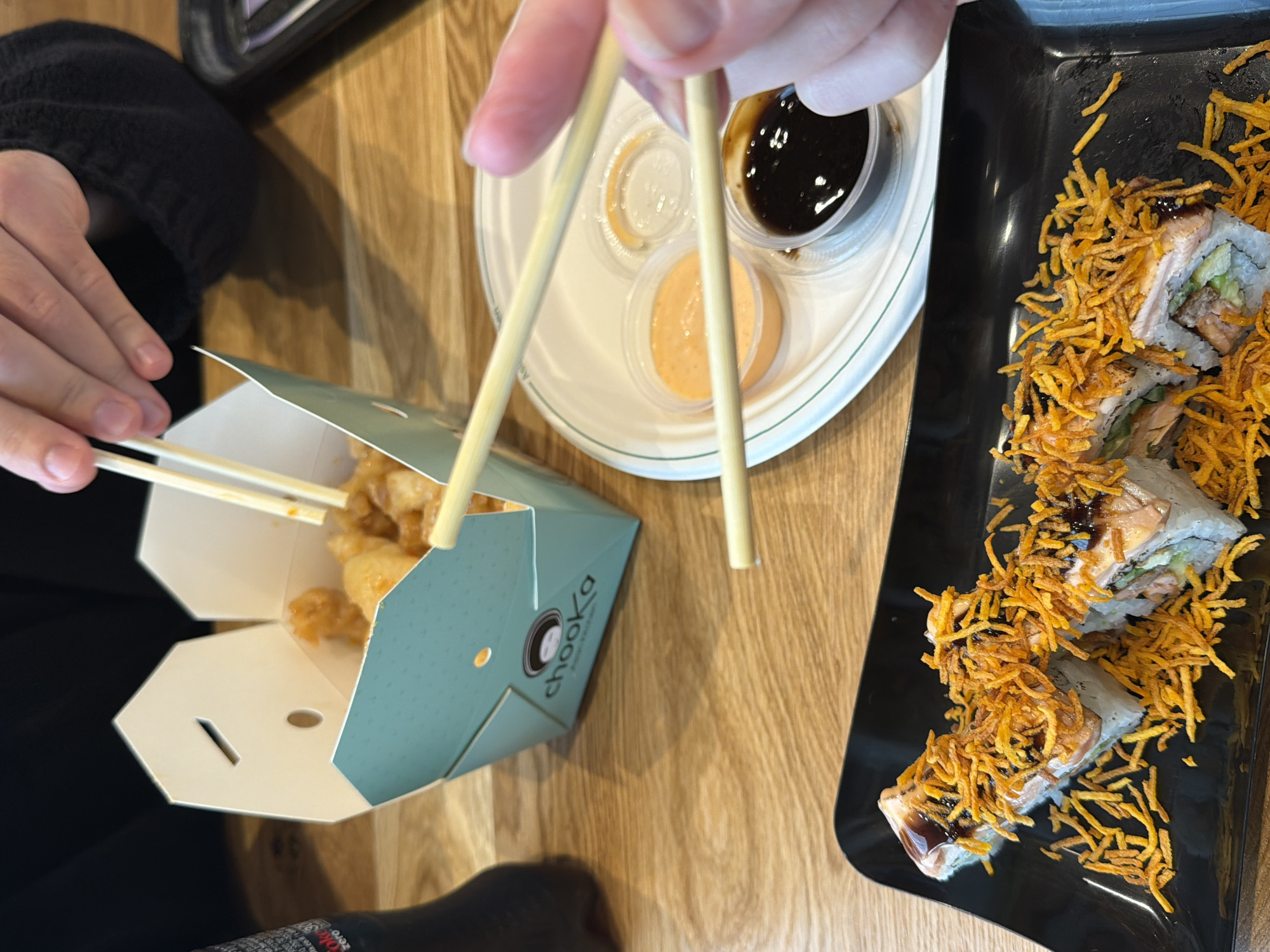 Here is a concise, forensic, matter-of-fact description of the image:
The image captures a dining scene, likely in a restaurant. A person uses chopsticks to pick up a piece of food from a small, open cardboard container with the logo "chooka" visible on its side. The container appears to hold fried chicken. The "chooka" logo is associated with restaurants in Tel Aviv, Israel, thus suggesting the location.
A white plate holds three small containers with sauces: one appears to be clear, one is orange-colored, and the other is a dark sauce, likely soy sauce.
To the right of the frame, a black plate contains several pieces of sushi rolls. The sushi is topped with thin, fried potato sticks and a dark sauce. The surface beneath the items is a light-colored, wooden-like tabletop.
The person holding the chopsticks is wearing a dark-colored top. The top of a container, maybe related to the order, is present in the upper left corner. The image shows an individual about to eat the takeout meal of friend chicken and sushi.
