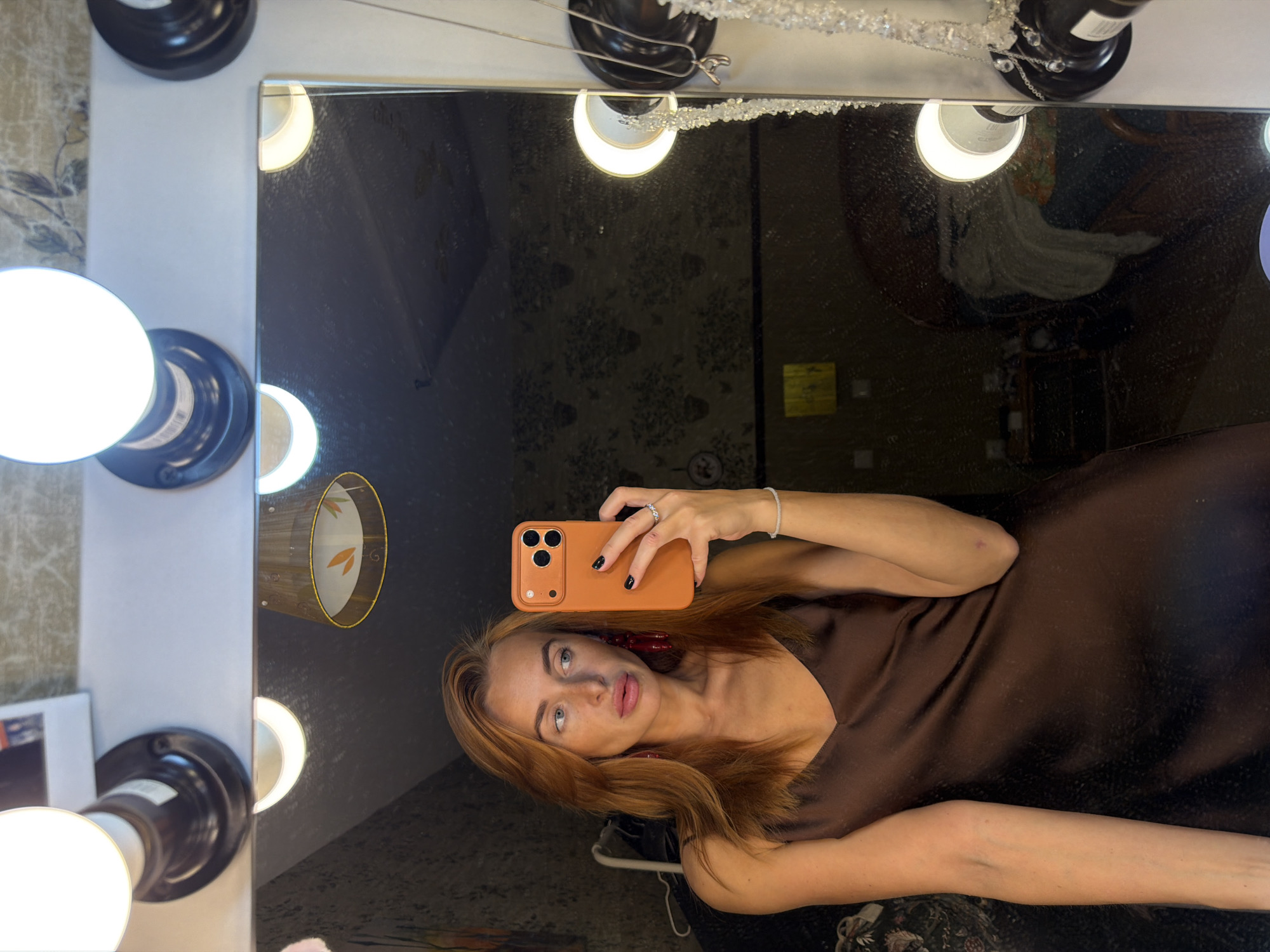 A woman with red hair takes a selfie in a mirror with vanity lights. The mirror reflects the interior of a room with a busy wallpaper pattern. A portion of the reflection suggests a seated figure. The setting could be a dressing room or similar space, potentially in Krasnoyarsk, Russia. The woman holds an orange smartphone, her hand adorned with a ring. She wears a brown satin dress.