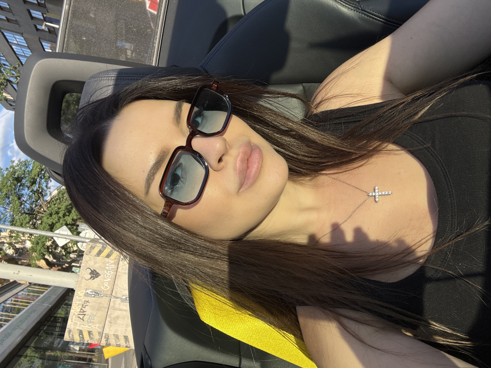 This is a close-up, low-angle, indoor-outdoor shot of a young woman taken in Prague, Czechia. She is lying back in what appears to be a car seat, possibly a convertible, with the top down. The woman has long, dark brown hair and is wearing sunglasses with thick, dark frames. She is also wearing a black tank top and a delicate silver necklace with a cross pendant. The sun is shining brightly, casting shadows on her face and chest, indicating it is daytime.
The background shows a mix of urban elements. To the left, a building with multiple windows can be seen, suggesting a city environment. Below the woman, a section of what looks like a street sign or advertisement with yellow and black hazard stripes is visible. There is also some graffiti on a wooden panel. Trees and blue sky are also present in the background, hinting at good weather.
The woman's face is turned slightly away from the camera, and her expression is relaxed, suggesting a leisurely or casual moment. The overall impression is one of enjoying a sunny day outdoors. There is text visible on the wooden panel which reads "GO VEGAN" along with a bull graphic. Other illegible text is also present.