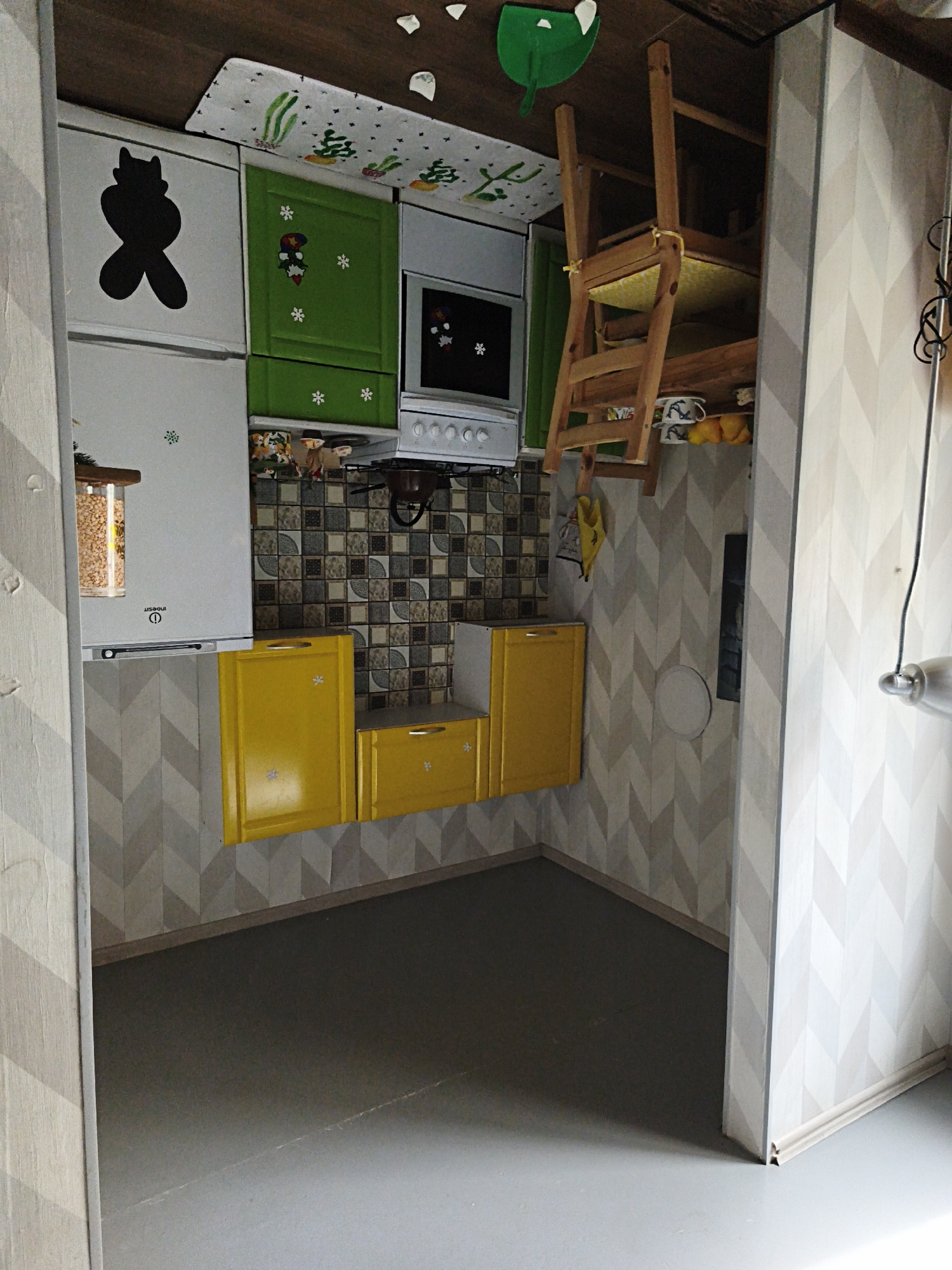 The image depicts an upside-down kitchen in what appears to be a staged exhibit or novelty installation. The room's contents are arranged in an inverted manner, with the floor essentially becoming the ceiling.
The kitchen features:
* Cabinets: Yellow lower cabinets and green upper cabinets, along with a white refrigerator.
* Appliances: A white oven and stovetop.
* Decor: A mat with cactus-like designs, and decorations with winter or festive themes (snowflakes, figures resembling Santa Claus) on the refrigerator and green cabinets.
* An upside-down wooden ladder with a cushion on the seat.
* Tiles: A section of tiled backsplash in a mosaic pattern.
The room's walls are covered in wallpaper with a geometric chevron pattern.
The floor is gray. The image gives no definitive indication that the location is Sevastopol, Ukraine, but one can infer it is situated in Sevastopol, Ukraine.