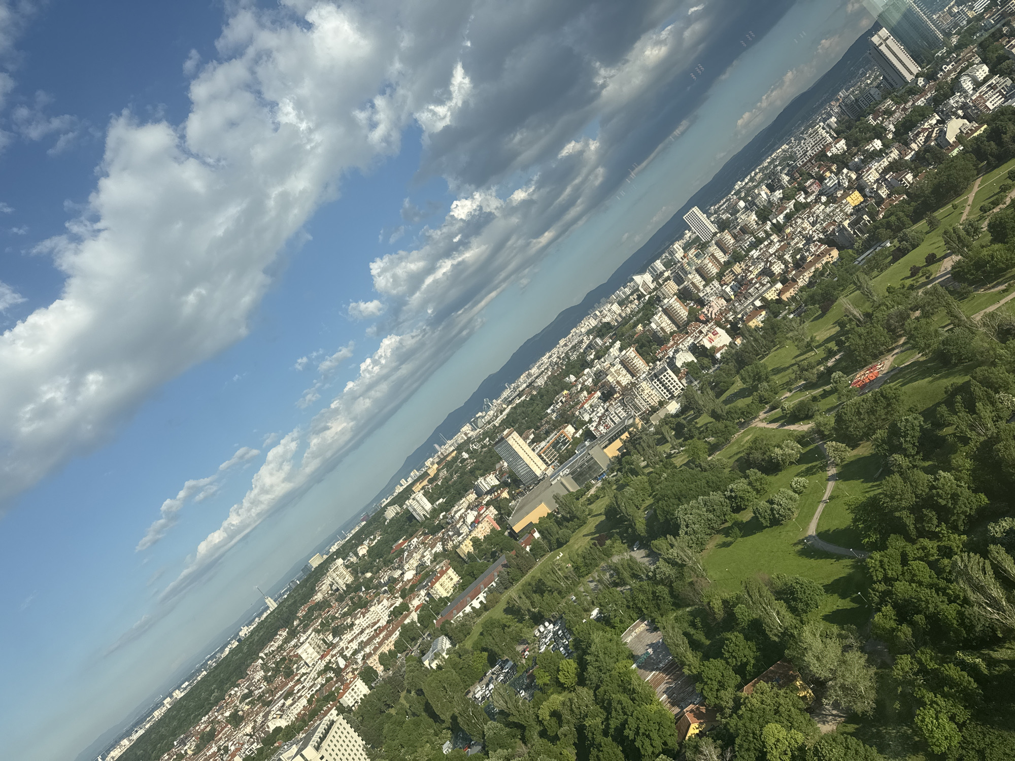 The image depicts an aerial view of Sofia, Bulgaria, captured on a day with mixed blue sky and cloud cover. The urban landscape is bisected diagonally by the viewpoint, showcasing a gradient from dense city blocks to green spaces. Buildings are predominantly low to mid-rise with a few taller structures interspersed. The city is nestled at the foot of a mountain range visible in the distance. Green spaces are abundant, featuring parks with walking paths and dense tree cover. The built environment displays a mix of residential and commercial properties. The light conditions suggest it is daytime, with shadows cast by clouds and buildings.