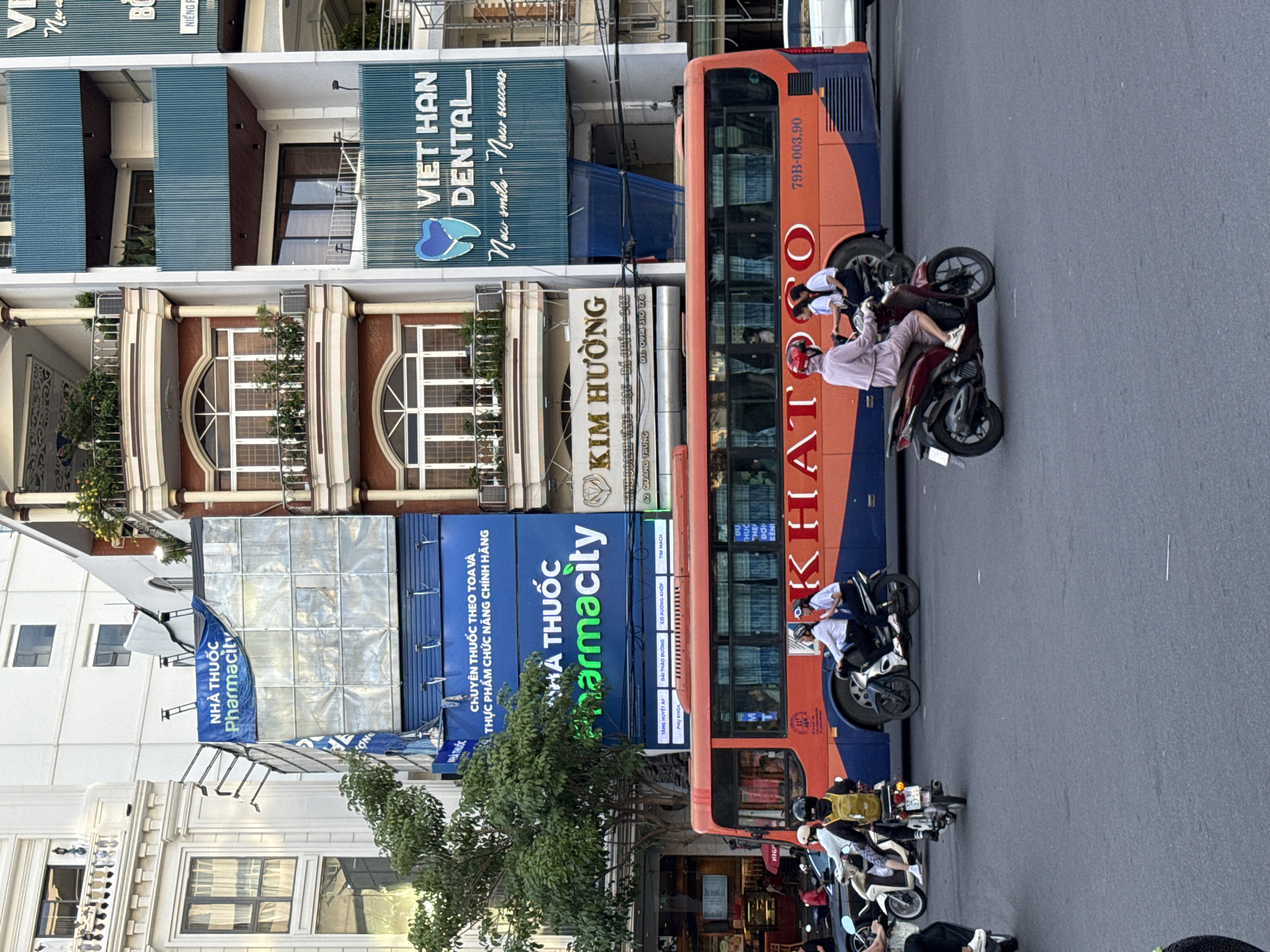 The image captures a street scene in Nha Trang, Vietnam, dominated by a large orange and blue bus. The bus is positioned centrally and displays the word "KHATOGO" in large red lettering along its side. Several motorbikes are present on the road, with riders and passengers. One motorbike in the foreground carries a person in a pink outfit. Behind the bus, buildings line the street, featuring various signage. Notably, a prominent sign for "NHÀ THUỐC Pharmacity" is visible, along with another for "VIET HAN DENTAL." The environment appears to be a busy urban area, with traffic and pedestrians. The lighting suggests it is daytime.