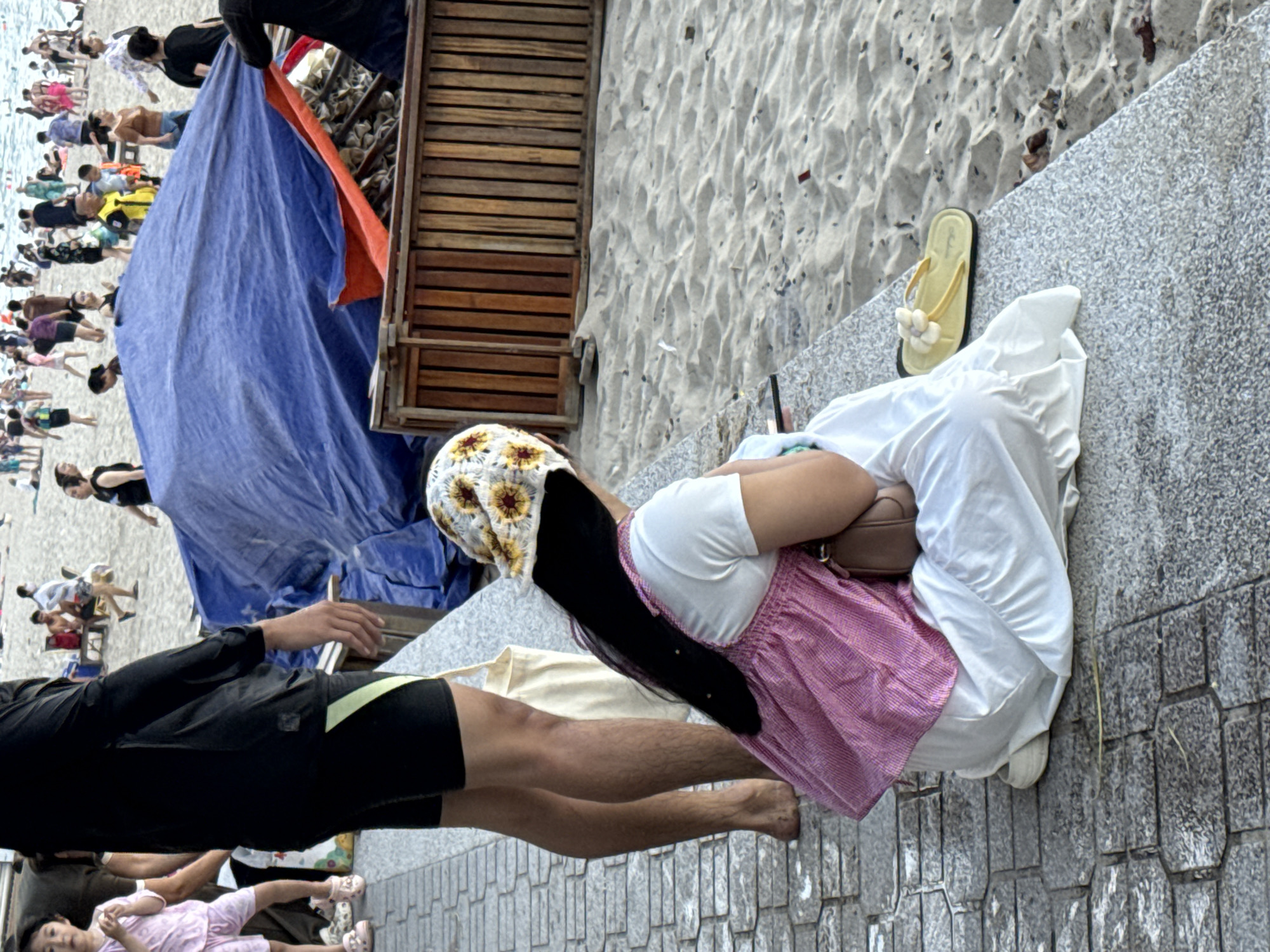 The image depicts a crowded beach scene in Nha Trang, Vietnam, with a focus on a woman sitting on a paved area adjacent to the sand, facing away from the camera. She is wearing a white t-shirt and a pink patterned dress, with a white and yellow sunflower-patterned headscarf. Her long black hair is visible. To her left, a person in black shorts and a black long-sleeved shirt stands with their legs visible, and a cream-colored tote bag hangs from their shoulder. In the background, numerous people are scattered across the sandy beach and in the water, engaged in various activities. A large blue tarp is spread out on the sand, partially covering wooden structures. The environment is characterized by sand, paved walkways, and the sea.