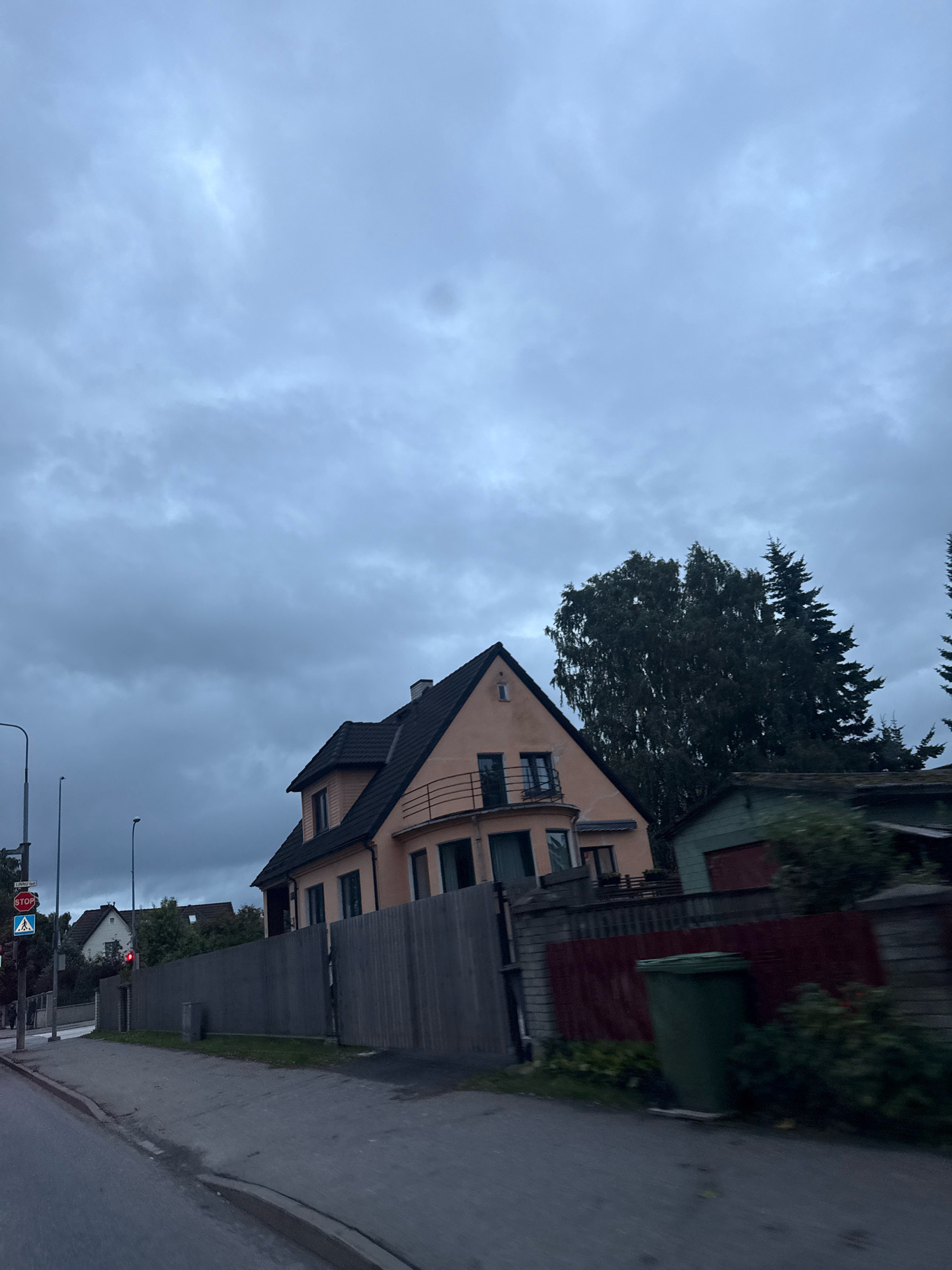 The image shows a street scene in Tallinn, Estonia, on a cloudy day. In the foreground, there is a paved road and sidewalk. A tall wooden fence runs along the right side of the road, separating it from a property with a peach-colored house. The house has a dark roof and multiple windows. To the right of the house, there is a green trash can and some overgrown vegetation. In the background, there are more houses and trees. A traffic light with a red signal is visible on the left side of the image, along with a STOP sign and a pedestrian crossing sign. The sky is overcast with thick, grey clouds.