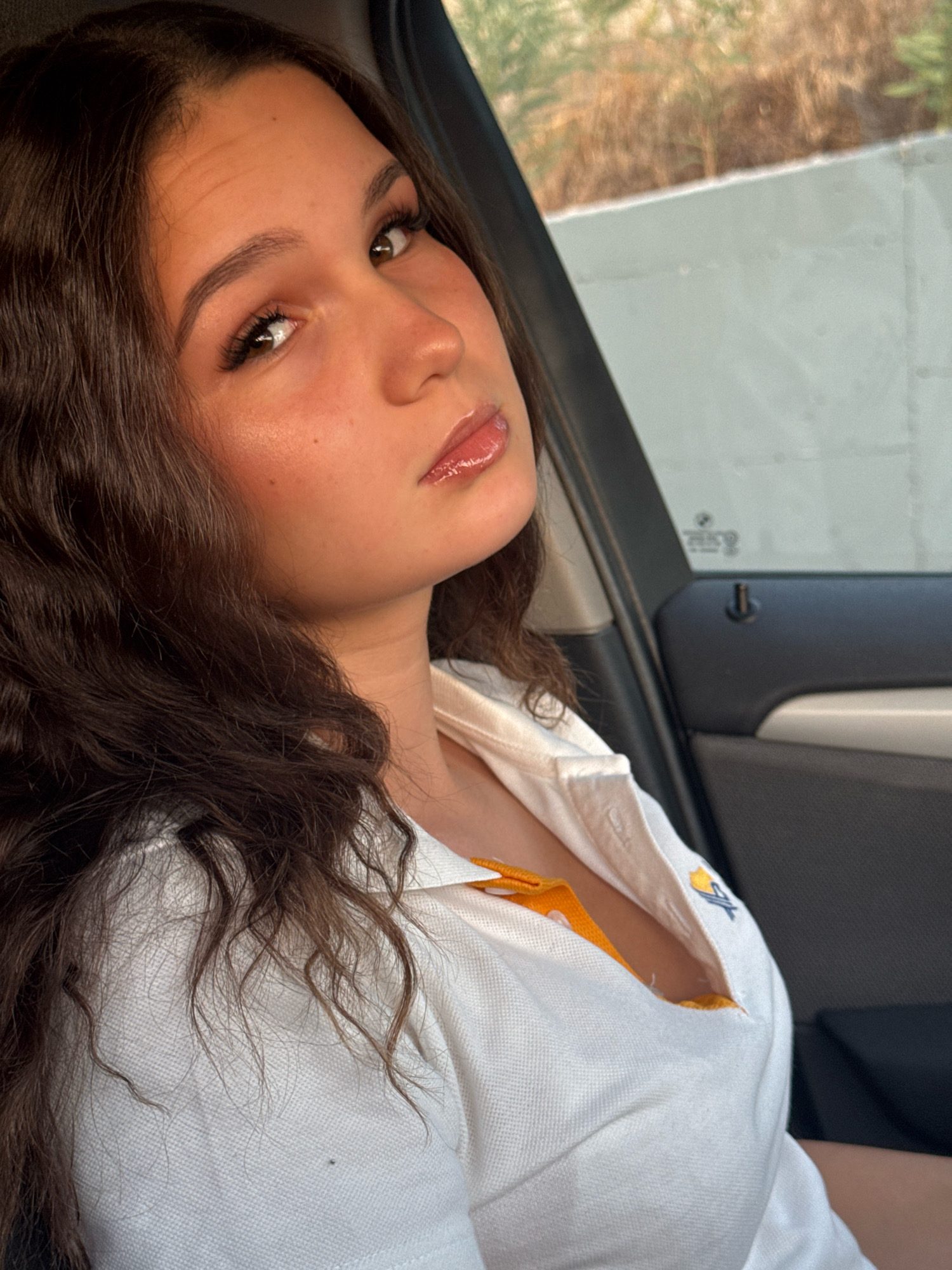 A young woman with long, dark, wavy hair and brown eyes is seated in a car, looking towards the camera. She is wearing a white collared shirt with orange trim visible underneath, and appears to be wearing makeup. The interior of the car, including the door panel and window, is visible. Outside the car window, a blurred background of foliage and a light-colored wall can be seen. The scene is likely captured during daylight. The location is Germasogeia Municipality, Cyprus.