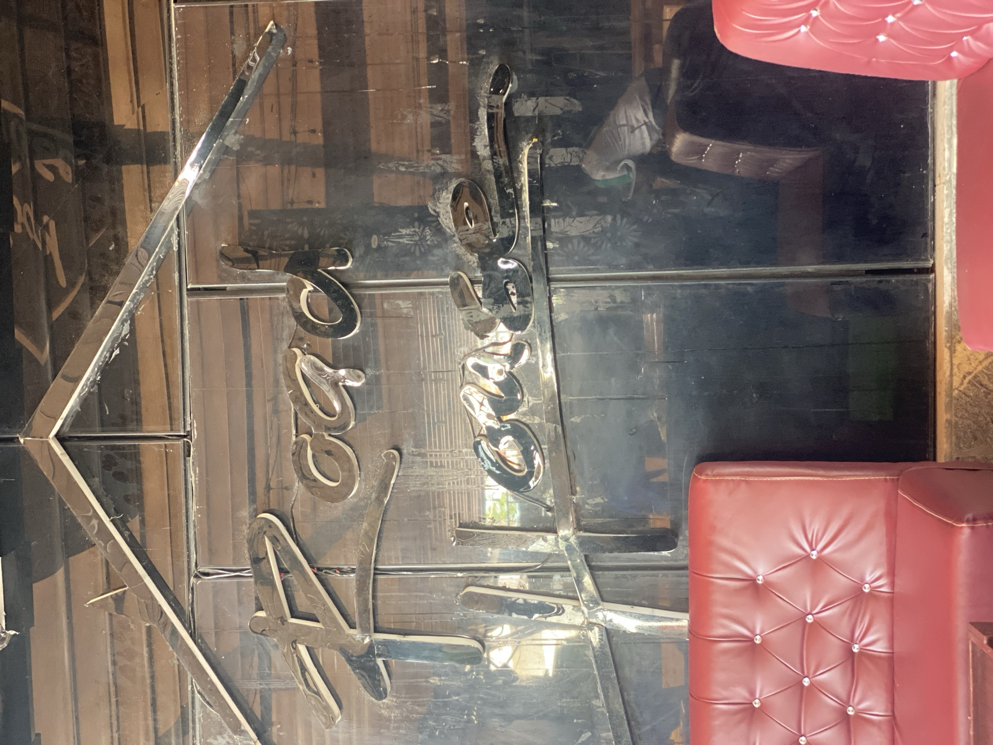 This is an eye-level, medium shot of a polished, reflective surface with chrome lettering that spells out "Roga Lounge". The lettering is stylized and has a vintage feel, with the "R" being particularly elaborate. The reflective surface is dark, possibly black or a very dark mirrored material, and shows reflections of the surrounding environment.
In the foreground, there are sections of a red tufted sofa with diamond-shaped tufting and clear jewel accents. The sofa appears to be made of leather or a similar material. To the right of the frame, another sofa, this one appears to be a dark brown color, is visible.
The background behind the lettering is a paneled wooden surface. Reflections in the dark surface include what appears to be outdoor greenery and sunlight, suggesting the location is in a commercial establishment. In the reflection, there is also a partial view of a person wearing a grey t-shirt.
The overall impression is of a lounge or bar interior, with a focus on the establishment's name displayed prominently. The location is identified as Howayi, Nigeria.