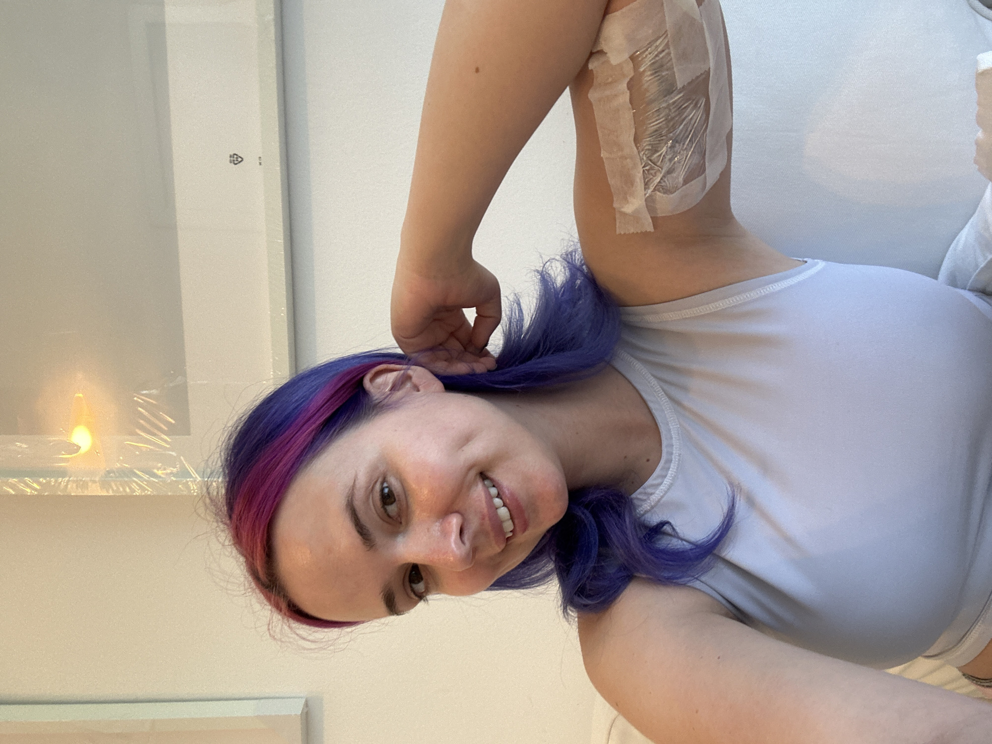 A medium close-up portrait shows a young woman with a warm smile, looking directly at the camera. Her shoulder-length hair is dyed a vibrant purple with pink or magenta highlights. She wears a light-colored (possibly white or light grey) sleeveless top.
On her upper right arm, near her shoulder, a medical dressing is visible. It consists of white adhesive tape securing a layer of clear plastic film over an area of her skin, suggesting a recent procedure or injury.
The background is simple and light-colored, indicating an indoor setting. To the woman's left, a large, rectangular white object, possibly a framed item or panel, is wrapped in clear plastic. On this object, a black recycling symbol (three arrows forming a triangle with the number "04" inside) and the text "LDPE" are visible. Further left, a soft, warm light, resembling a candle flame or small lamp, reflects on the plastic wrapping. The woman's expression conveys happiness and contentment.