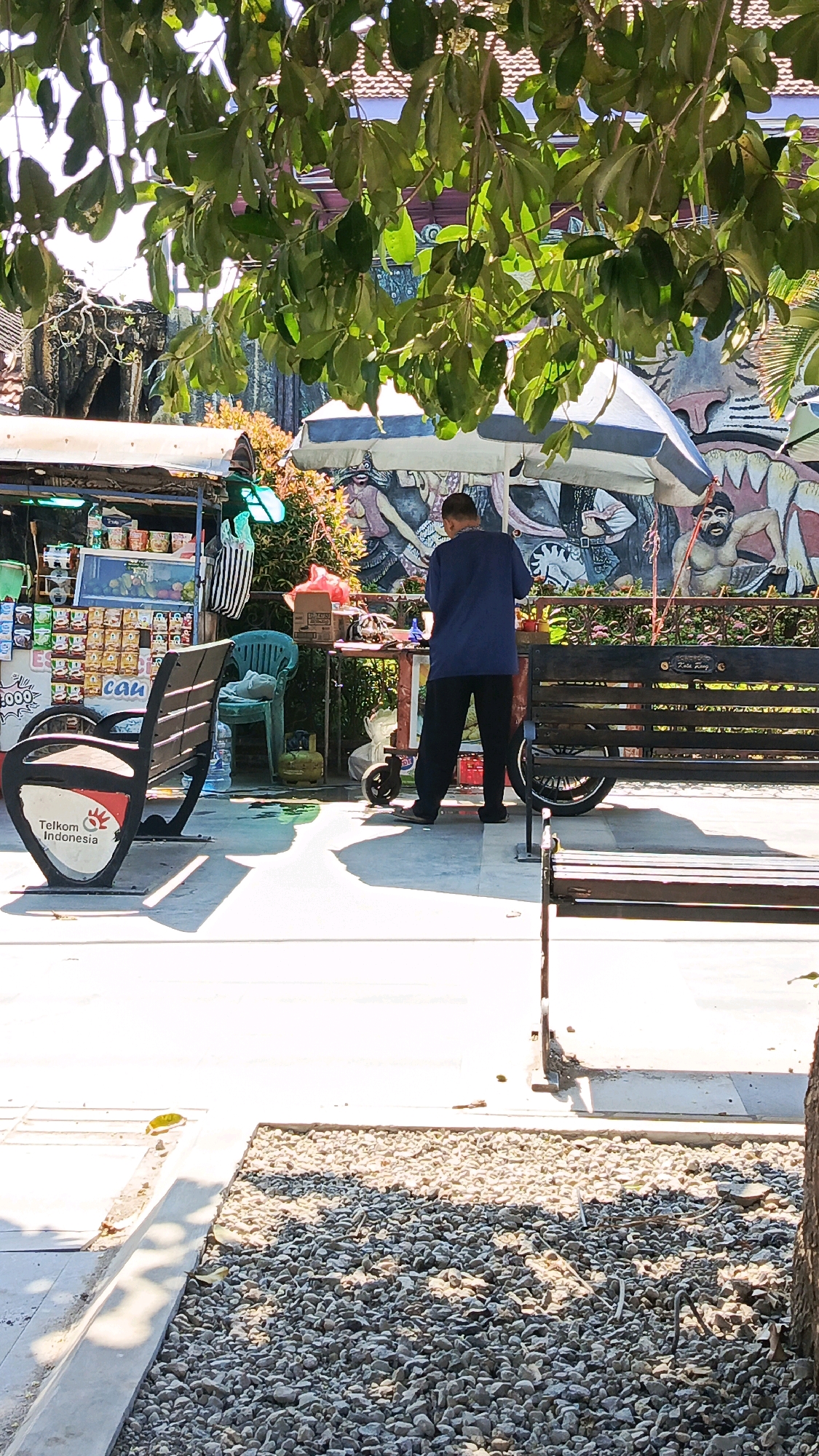 In Ponorogo, Indonesia, a man stands behind a food stall under the shade of lush green trees. The stall, adorned with colorful snacks and beverages, is positioned in front of a vibrant mural depicting traditional Indonesian figures. To the left, a bench with a "Telkom Indonesia" logo invites patrons to rest, while another wooden bench sits to the right. The ground is a mix of smooth concrete and rough gravel, suggesting an outdoor public space, possibly a park or pedestrian walkway. Sunlight casts long shadows, indicating a bright, sunny day.