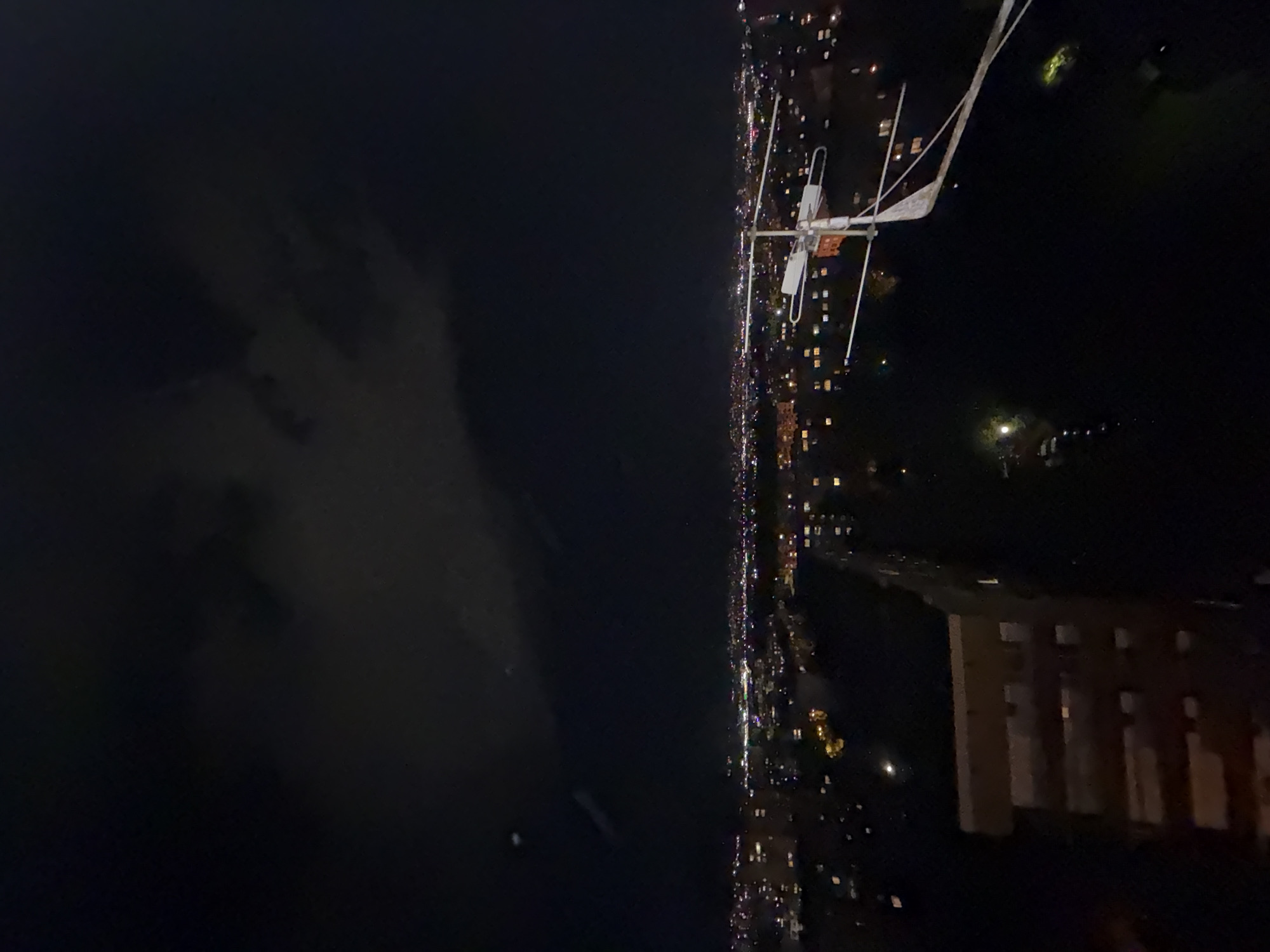 A vertical photograph captures a sweeping night view over a city, with a distinct horizon line dividing the dark sky from the illuminated urban landscape.
In the upper-left portion of the image, the dark night sky features soft, diffuse cloud formations that are subtly visible against the darkness, catching ambient light from below. The rest of the upper frame is a deep, unlit expanse.
Dominating the mid-right side of the image is a prominent, intricate white metallic structure, likely an antenna or weather monitoring equipment. This apparatus consists of multiple horizontal and vertical bars, with what appears to be white fabric or plastic elements attached to some parts, possibly covers or flags.
Below the horizon line, the city stretches out with countless twinkling lights, forming a vibrant tapestry that extends horizontally across the lower half of the frame and recedes into the distance. These lights delineate streets, buildings, and residential areas, creating a bustling impression against the dark ground. On the lower-right side, a large, dark rectangular building with several illuminated windows is discernible, adding structural detail to the urban environment.
There are no people or specific activities depicted in the scene, which offers a static, serene yet expansive vista of a city at night under a partly cloudy sky. The general setting is an urban landscape, identified as Sofia, Bulgaria, though no specific landmarks are visible in the image. No visible text is present.