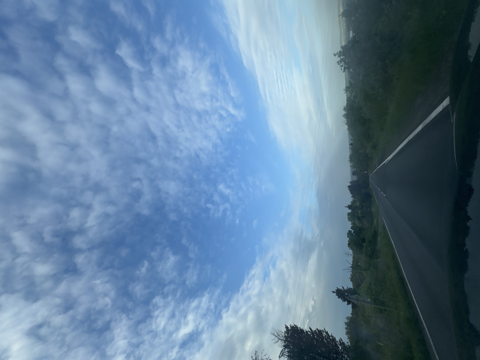 The image presents a view from a vehicle on a road, likely in a rural or semi-rural area based on the surrounding vegetation. The sky occupies a significant portion of the image, displaying a mix of clear blue areas and scattered clouds. The road stretches into the distance, with trees and greenery lining its sides. Power lines are visible crossing the sky near the road. The reflection of the scene can be seen on the car's surface. There is no definitive evidence of the location being in Nesebar, Bulgaria.