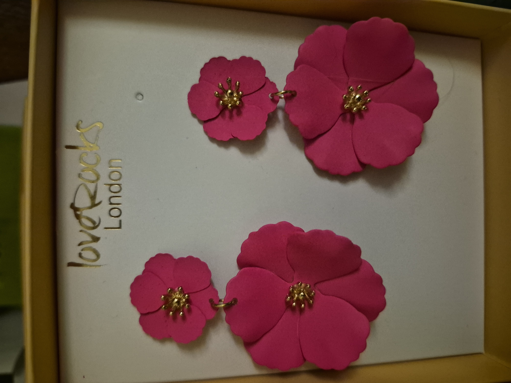 A pair of fuchsia-colored flower-shaped earrings are displayed in a golden box. The earrings are layered, with a smaller flower hanging above a larger flower, both featuring a gold-colored center. The brand name "loveRocks London" is printed in gold lettering on the white background of the box. The image is shot from a top-down perspective, focusing on the earrings.