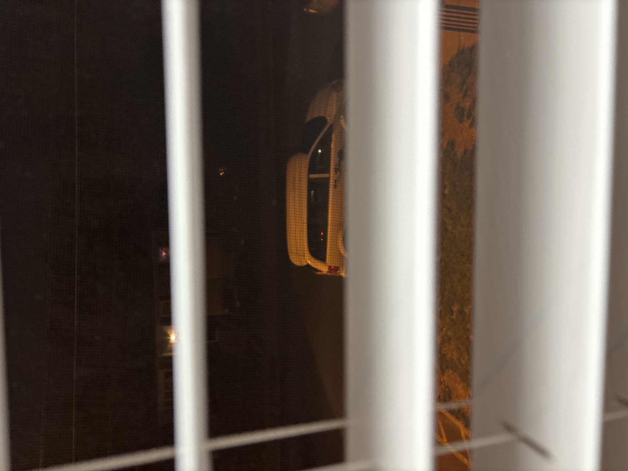 The image depicts a nighttime scene viewed through a window partially obscured by white vertical blinds. The primary subject is a white car, possibly a sedan or coupe, seen parked outside through the gaps in the blinds. Its rear section is most prominent, revealing a red taillight glowing faintly. Reflections from inside the car or a nearby light source are also visible on its surface.
The setting is dark, indicating it is night or late evening. The background is largely undifferentiated black, with a few faint light sources visible in the distance, possibly from other buildings or streetlights. To the right of the car, a textured surface, which appears to be pavement or a driveway, can be seen between the blind slats. The image itself is oriented vertically, suggesting it has been rotated 90 degrees clockwise from its original capture orientation. There are no people visible, nor is any discernible text present in the image. The scene is located in Franklin Township, United States of America.