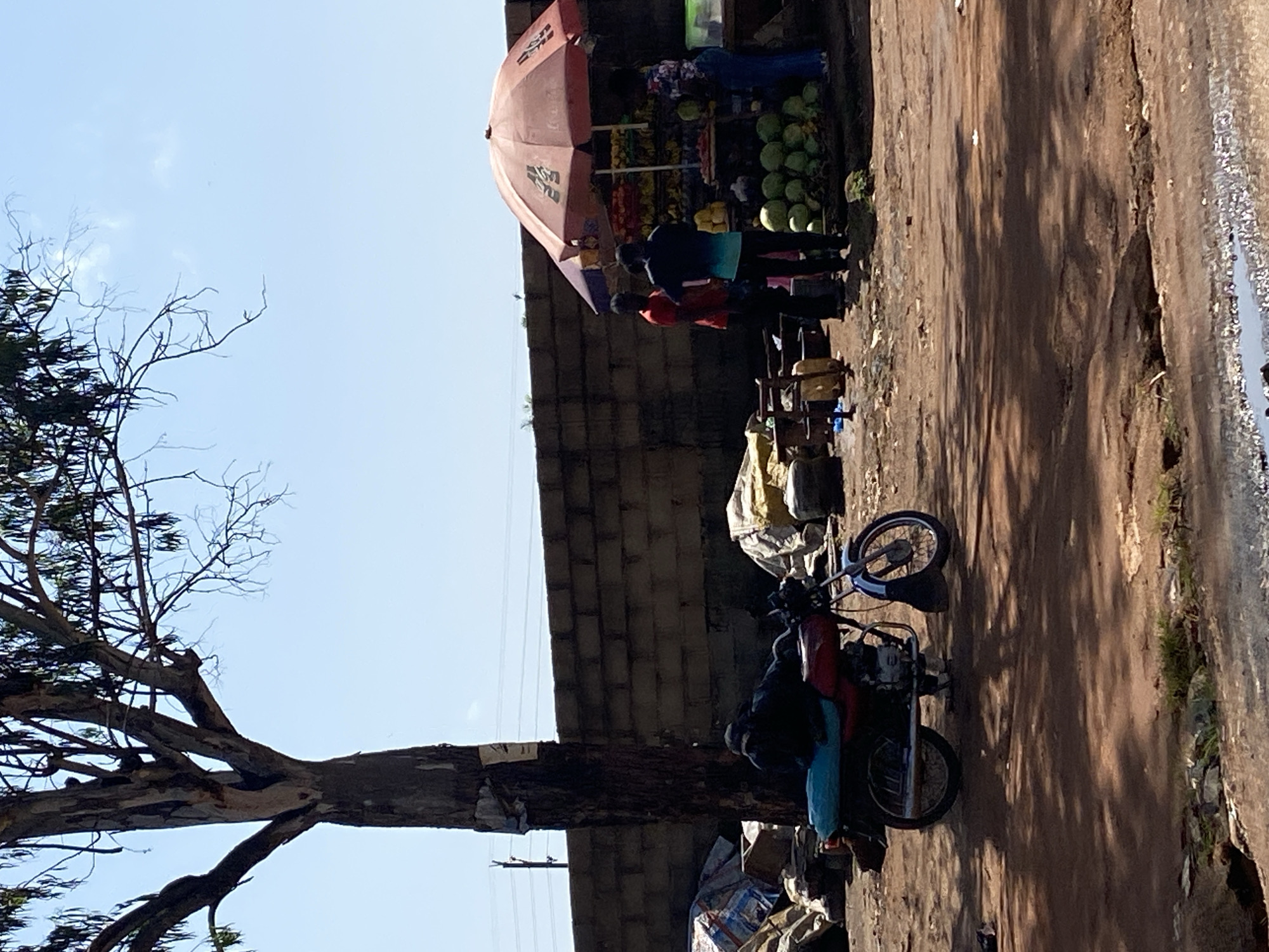 In Gura Top, Nigeria, a scene unfolds on a dusty, sun-drenched street. A motorcycle, partially obscured by shadows, is parked in the foreground. It is a red and blue model with a turquoise seat. A person, dressed in dark clothing, is seated on the motorcycle, appearing to rest or be asleep. To the left, a large tree with sparse leaves casts a shadow across the ground, its bark rough and textured. Behind the motorcycle, a pile of assorted goods, including sacks and crates, are visible. Further back, a fruit stand operates under a large, orange umbrella. Several people are gathered around the stand, some interacting with the vendor. The fruit stand displays a variety of produce, including watermelons, bananas, and other fruits and vegetables. A brick wall forms a backdrop to the scene, with power lines visible against the clear blue sky. The ground is uneven, covered in dirt and scattered debris, indicative of a typical street market environment.