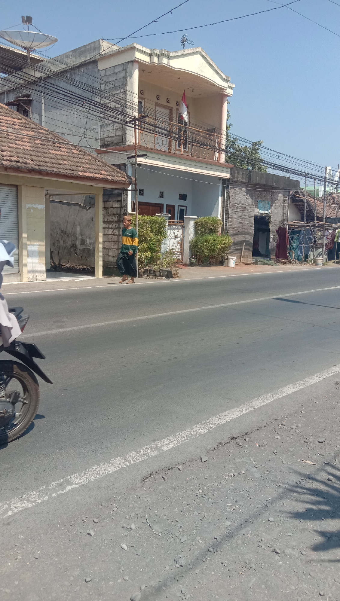 The image shows a street scene in Karang Anyar, Indonesia. A man in a yellow and green striped shirt and dark pants is walking on the sidewalk next to the road. Behind him are several buildings, including a multi-story house with a balcony and an Indonesian flag hanging from it. There are also some smaller buildings and a satellite dish visible. The road is paved and has a white line marking the lane. A motorcycle is partially visible on the left side of the image. The sky is clear and blue.