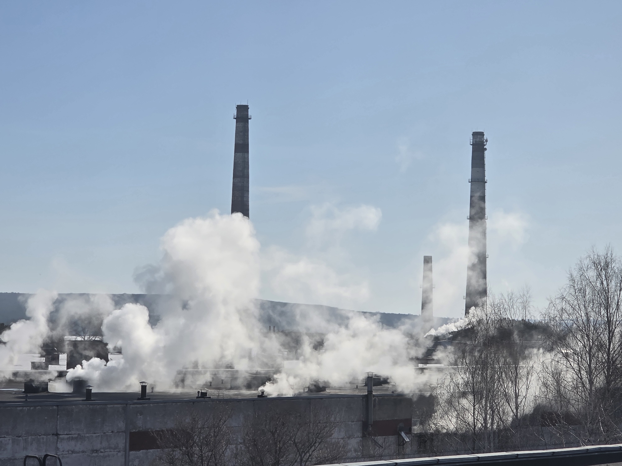 The image depicts an industrial scene, presumably in or near the city of Chaikovsky, Russia, given the prompt. There are at least three tall industrial chimneys or smokestacks. Two are prominently visible, while a third, shorter one is partially obscured in the background. Plumes of white smoke or steam are being emitted from the structures on the ground. Some smoke also appears to be coming from the chimneys. The lower portions of industrial buildings are visible. In the foreground, there are bare trees and a concrete wall. The sky is pale blue.