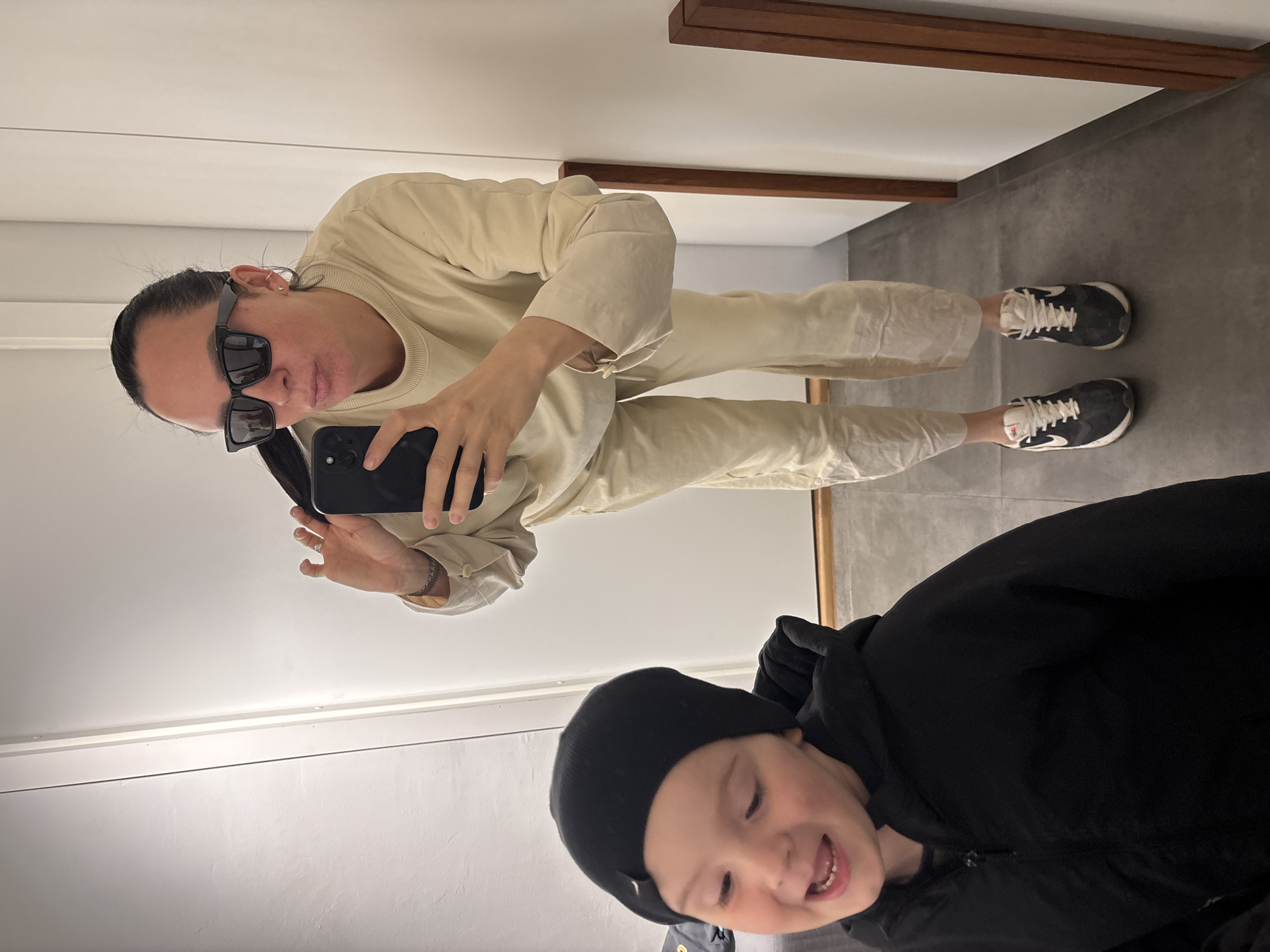 A selfie taken in a room with gray flooring and white walls shows a woman wearing black sunglasses, a beige top, and matching pants, standing with her feet visible in black and white shoes. She is holding a smartphone up to take the picture. A child wearing a black hat and jacket is partially visible in the lower right corner of the frame. There is a wooden beam detail near the ceiling. The location is Chaikovsky, Russia.