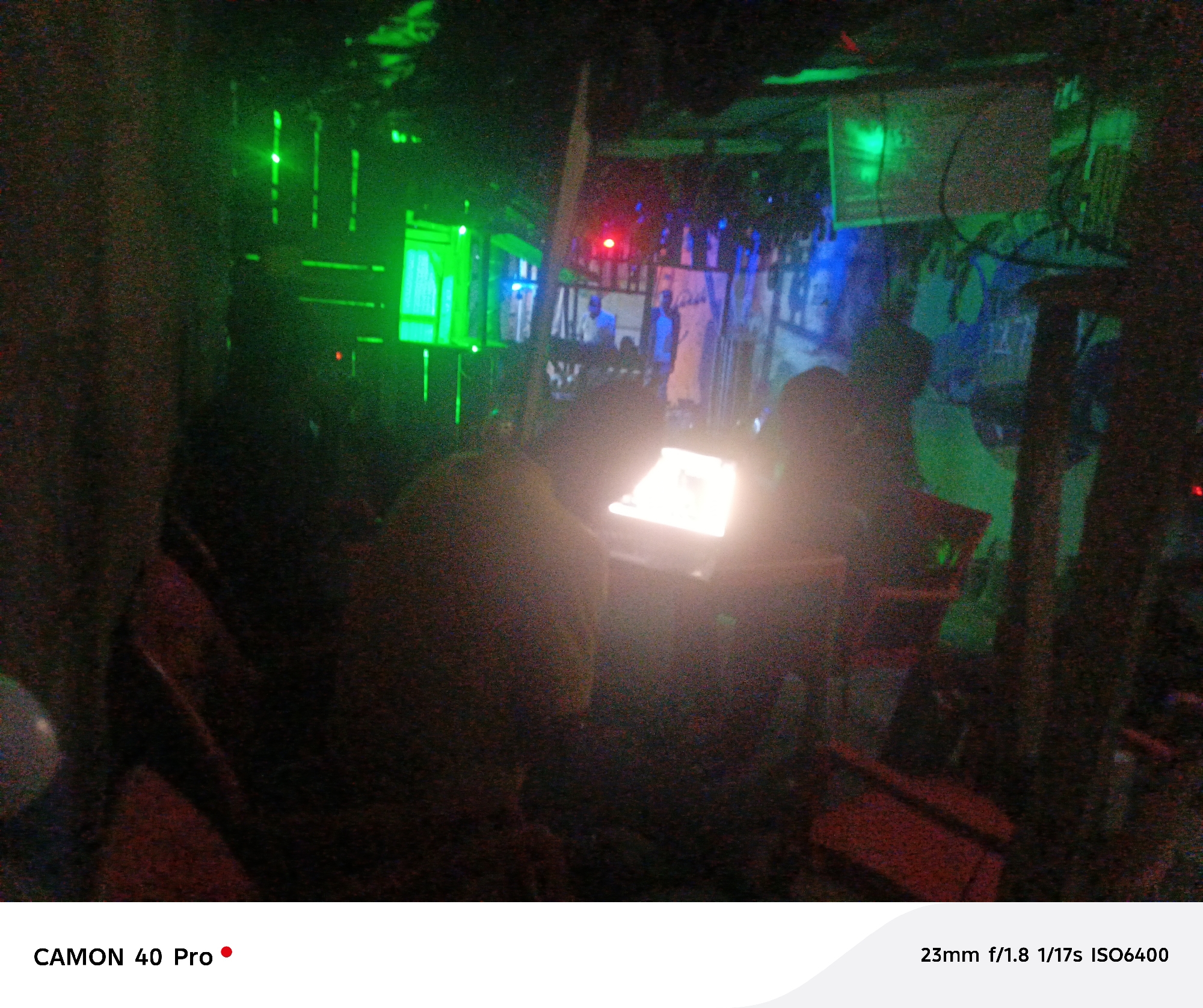 The image depicts an interior scene with low lighting, possibly in Jos, Nigeria. Two individuals are visible, seated at a table with a bright screen in front of them, likely a laptop or monitor. The background features various elements including green and blue neon lights, posters or murals, and structures that appear to be part of the establishment's decor. The overall atmosphere suggests a gaming center or internet cafe. The foreground includes chairs and other furniture, emphasizing the seated perspective of the scene.