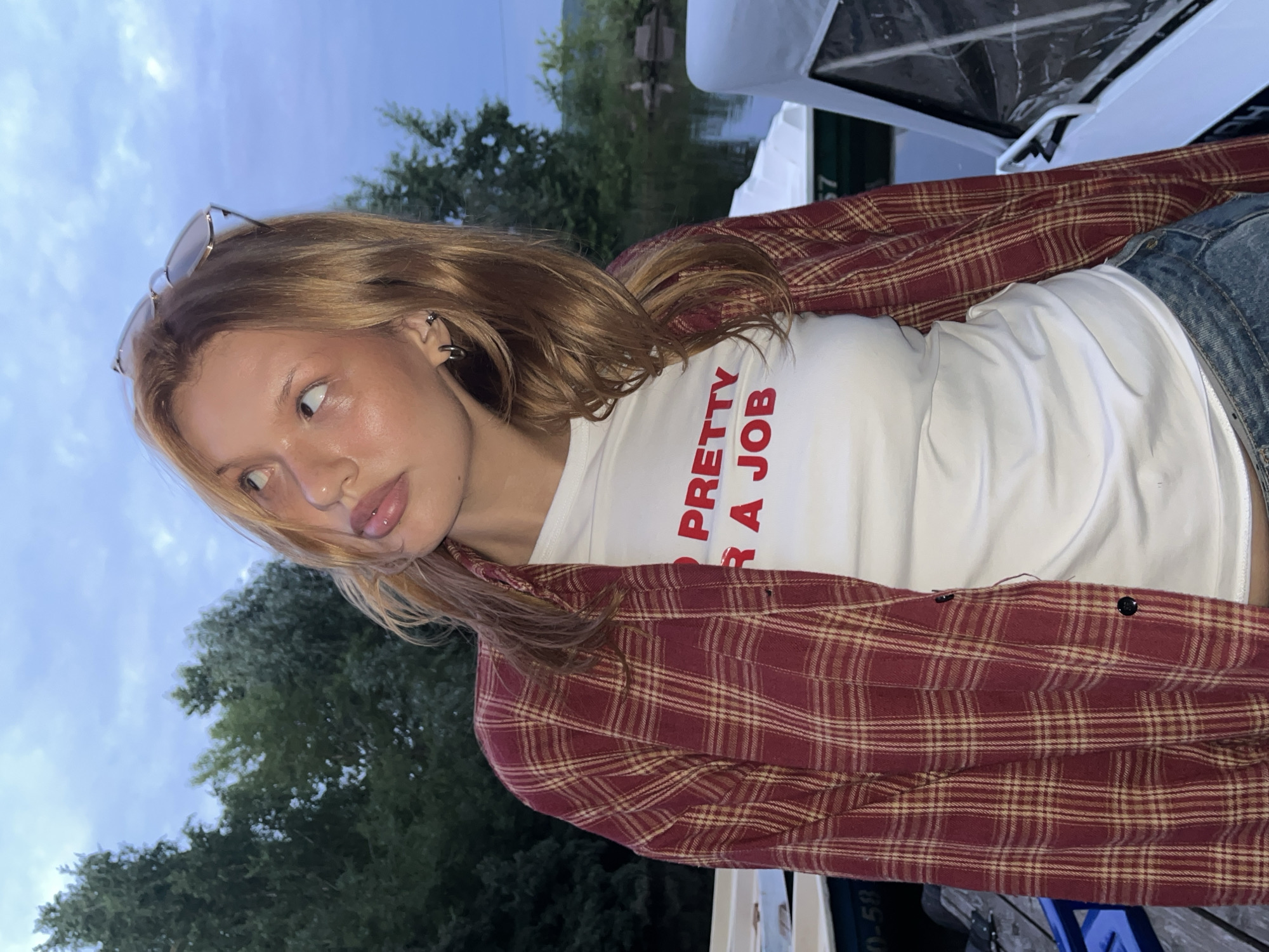 An outdoor medium shot captures a young woman with blonde hair and fair skin looking upwards and to her left. She is wearing a white t-shirt with red lettering that reads "PRETTY FOR A JOB", and an open red and tan plaid shirt over it. She has hoop earrings in both ears and sunglasses perched on top of her head. The background shows a body of water, possibly a river or lake, with several boats docked. Lush green trees line the opposite bank. The sky is overcast with a bluish-grey hue, suggesting it is either early morning or late evening. The overall setting appears to be a waterfront area within the city of Chaikovsky, Russia, likely a park or marina, given the presence of boats and the natural landscape.