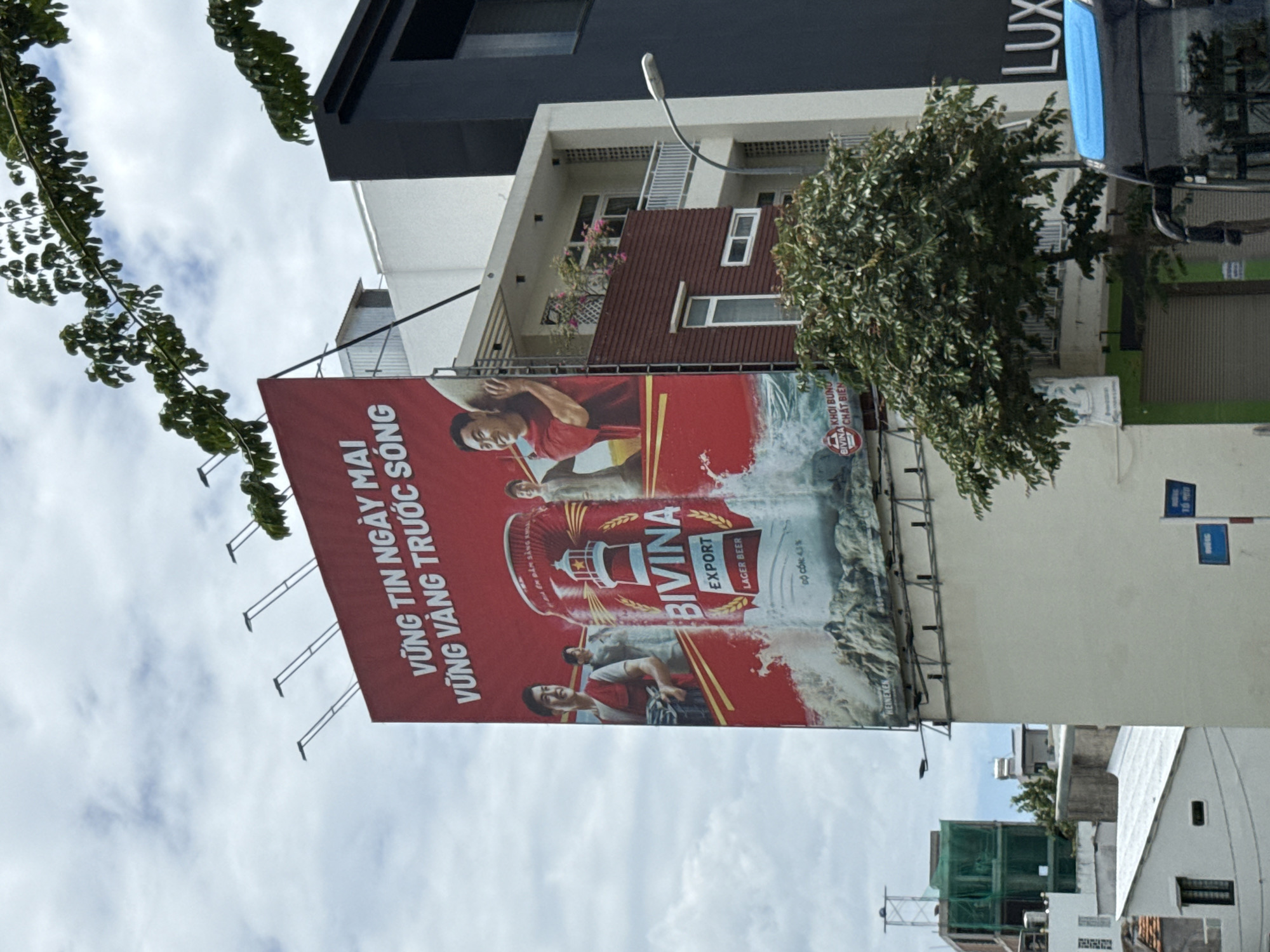 This is a street-level, full-shot photograph taken in Nha Trang, Vietnam. The image is dominated by a large billboard advertising "Bivina" beer. The billboard features images of three men, a large can of beer, and Vietnamese text. Below the billboard, part of a black building with "LUX" written on it is visible. A street lamp is positioned in front of the buildings. In the foreground, the top of a blue bus is visible. A large green tree partially obstructs the view of the billboard and buildings. The sky above is cloudy.