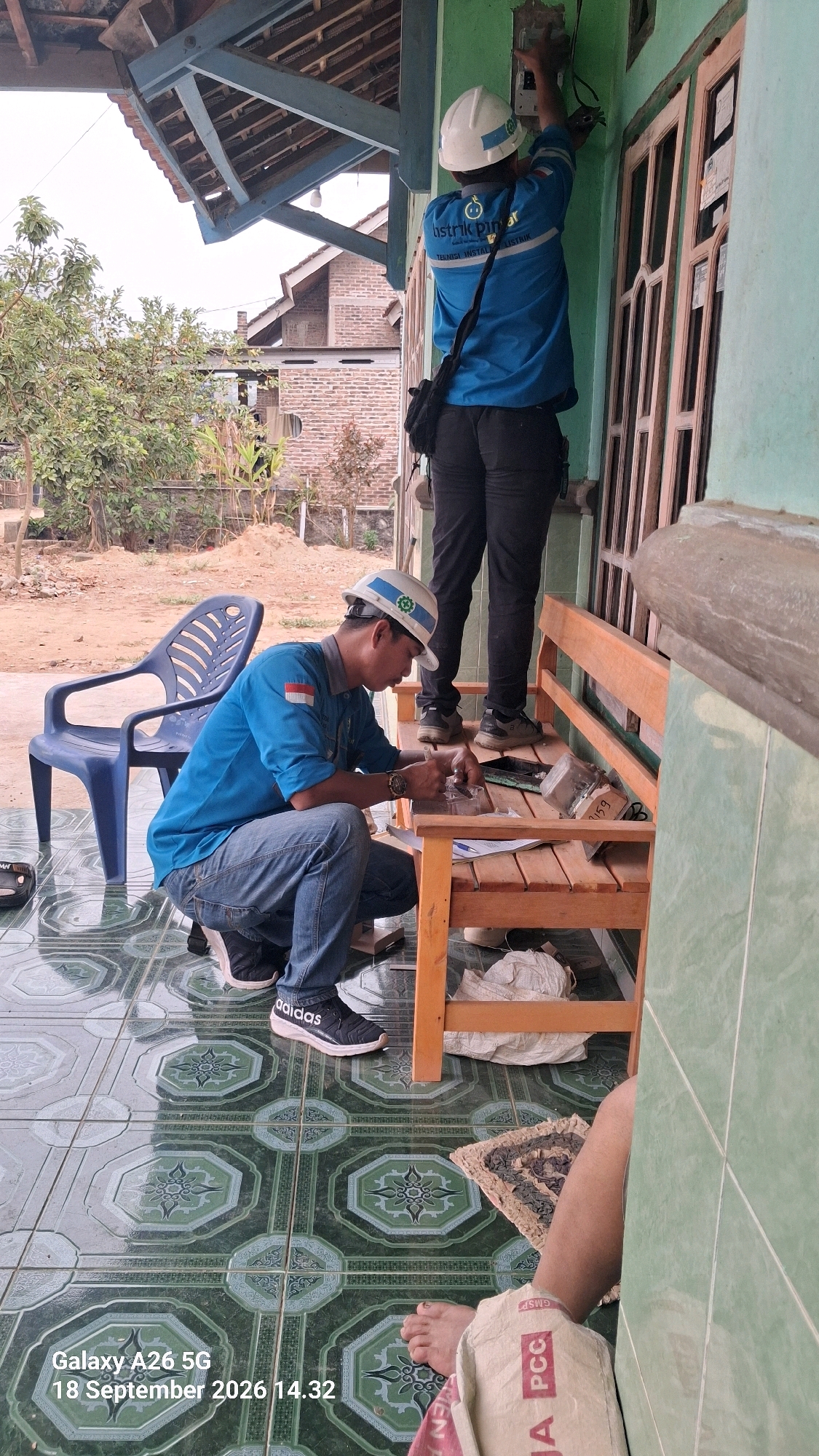 Two male individuals in hard hats are present at a residential property. One individual, wearing a blue uniform shirt with "Listrik PLN" branding, is standing and appears to be working on an electrical meterbox affixed to the wall of a light green building. He is also carrying a black shoulder bag. The second individual, also in a blue uniform shirt and hard hat, is crouching on a tiled porch, looking down at a wooden bench. He appears to be handling some small objects or tools. A blue plastic chair is visible on the porch, and a wooden bench is positioned in front of the building. The environment suggests an outdoor residential setting, possibly in the city of Karanganyar, Indonesia, with a tiled floor and some vegetation in the background. The timestamp "Galaxy A26 5G 18 September 2026 14.32" is overlaid on the image.