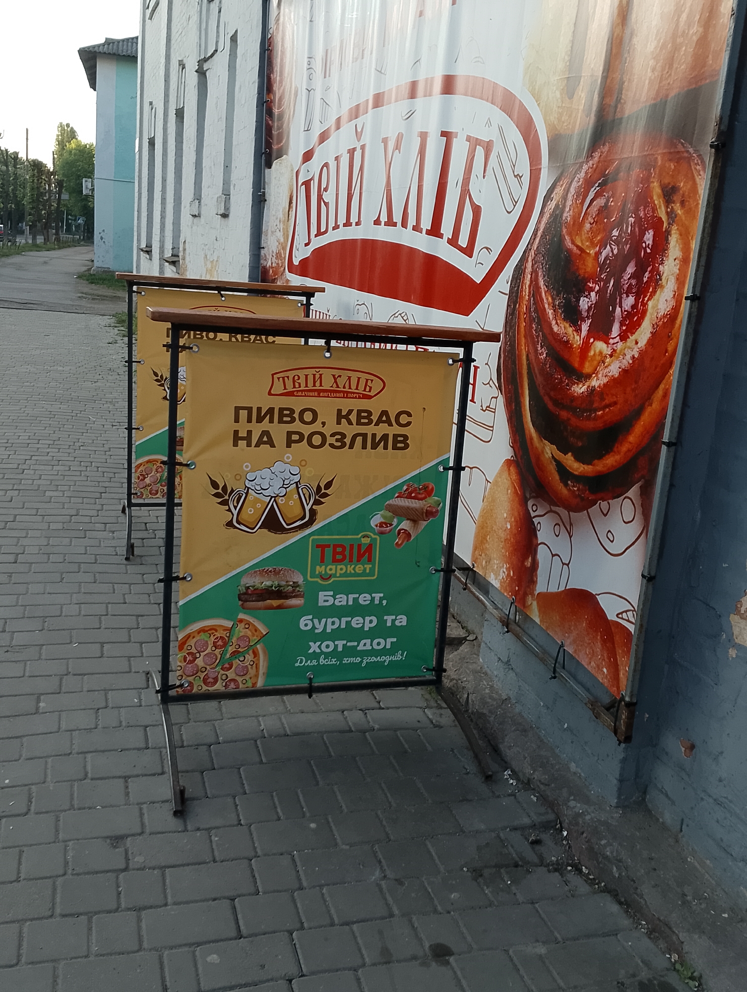 Here's a detailed description of the image:
**Setting:** The image appears to be taken on a sidewalk in an urban area, likely a city in Ukraine, judging by the Ukrainian text on the signage. The sidewalk is paved with gray interlocking bricks. Adjacent to the sidewalk is a building with a distressed, light-colored facade. In the background, there's a glimpse of a road and buildings, suggesting a residential or commercial area.
**Objects:**
* **Signage:** There are two prominent advertising signs. The first one closer to the viewer advertises "Твій Хліб" (Your Bread), beer, kvass (a traditional fermented beverage), baguettes, burgers, and hot dogs. It features images of these items along with the text. The second, larger sign displays an image of sweet bread.
* **Sign Holders:** The signs are held up by metal frames with wooden crossbars.
* **Building:** The building alongside the sidewalk appears to be constructed of brick, and painted in lighter shades, with signs of weathering and chipping paint.
* **Roadway and Buildings:** In the background, there's a paved road with other buildings across it, indicating the scene is in a populated area.
**Environment:** The lighting suggests it's either early morning or late afternoon, with soft shadows cast by the signs. The presence of trees in the background suggests some greenery in the surrounding area. The overall atmosphere appears to be quiet.
**Actions/Interactions:** No people are visible in the shot, so there are no direct actions or interactions. The signs are positioned to attract the attention of passersby.
