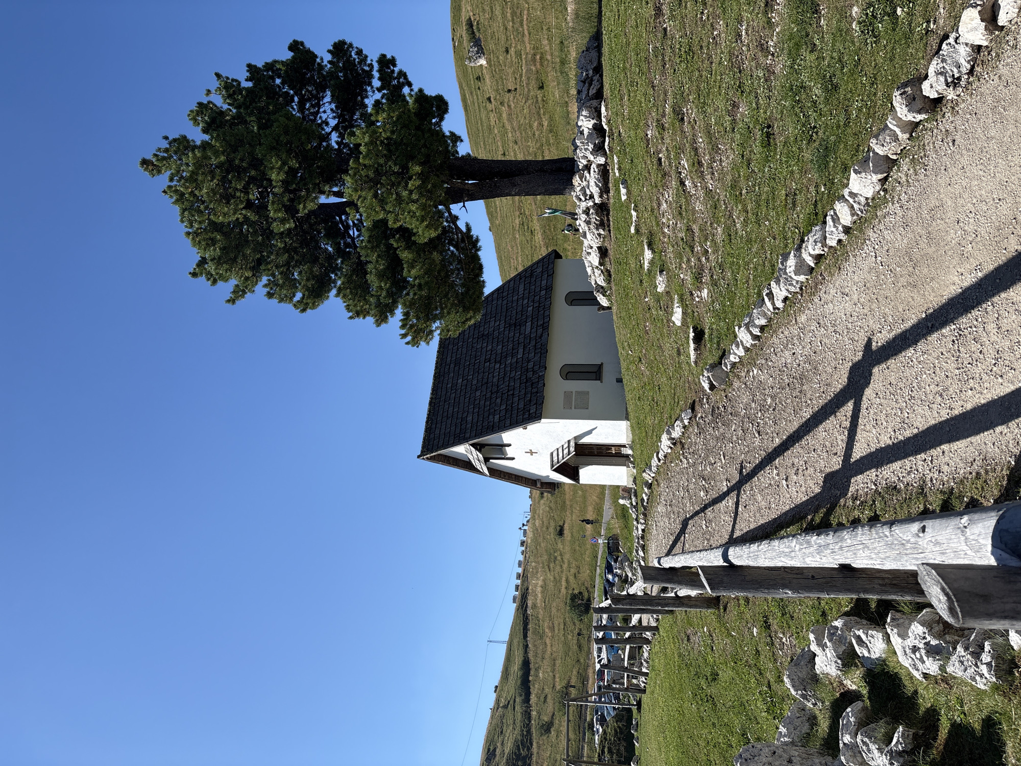 The image depicts a small white chapel with a dark, shingled roof, situated on a grassy hillside under a clear blue sky. A dirt path, lined with small white rocks, leads up to the chapel. To the right of the chapel, a large evergreen tree stands prominently. In the background, there is a parking lot with several cars, a wooden fence, and rolling hills covered in grass. The setting appears to be a rural or mountainous area. The location is identified as the Church of San Silvestro, Sëlva - Wolkenstein - Selva di Val Gardena, Italy.