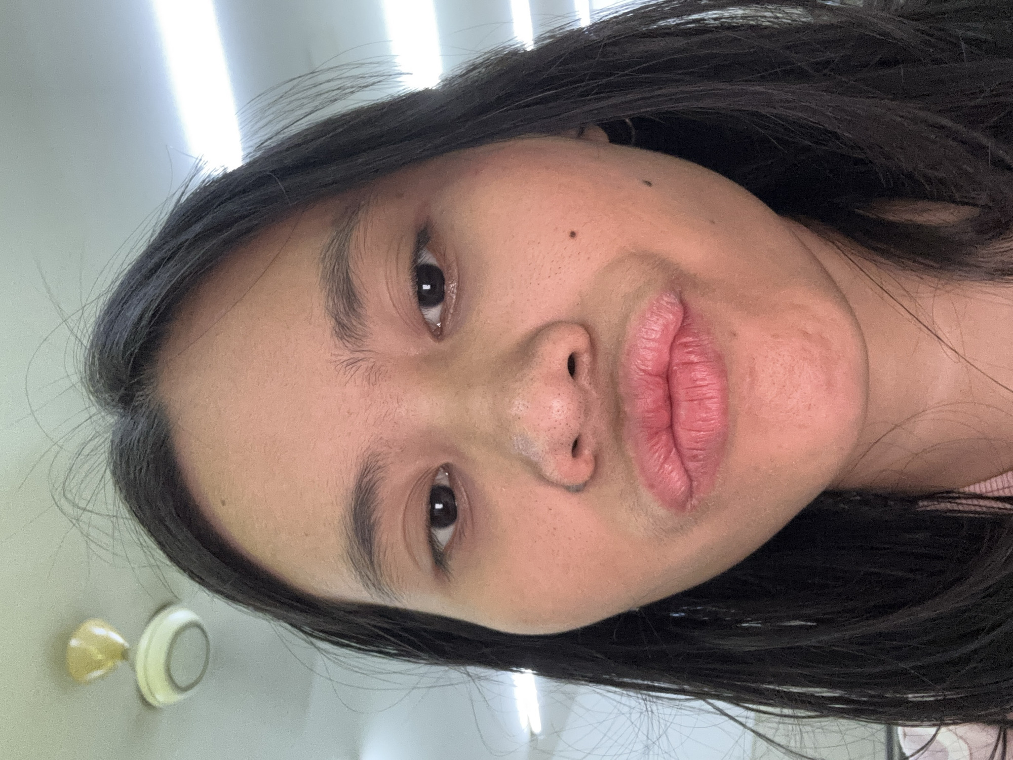 A young woman with long, dark hair is taking a selfie, puckering her lips towards the camera. She is indoors, with fluorescent lights and a ceiling fan visible in the background. The scene appears to be a brightly lit room. The woman has a mole on her right cheek.