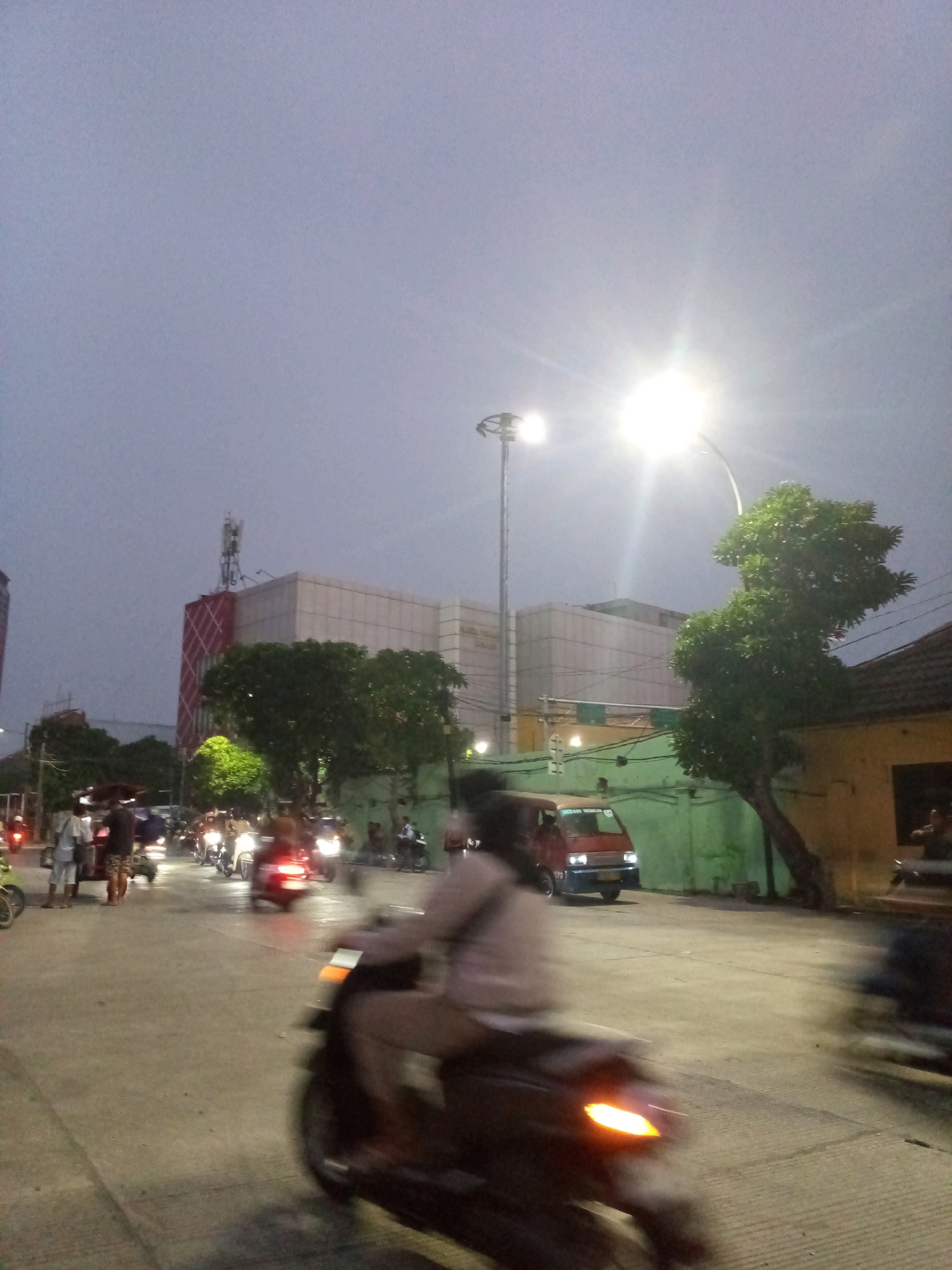 This is a long-exposure photograph taken at dusk in Bekasi, Indonesia, depicting a street scene with motion blur due to moving vehicles. The sky is overcast and a pale gray. A bright streetlamp in the upper right quadrant illuminates the scene, casting a strong glare. In the background, a modern, light-colored building with a red-patterned facade and a cell tower on top is visible. Adjacent to it is a long, light-colored building with what appears to be windows or facade elements. To the right of this, a green wall runs along the property line, with a yellow building beyond it, partly obscured by a tree with dense foliage. A red and blue van is parked on the street. Several motorcycles, their riders blurred by motion, are traveling along the asphalt road. Some pedestrians are also present on the left side of the road, appearing as indistinct figures. The foreground is dominated by a sharply blurred motorcycle moving from left to right, its headlight emitting a bright orange glow. The overall impression is one of a busy street at twilight in an urban environment.